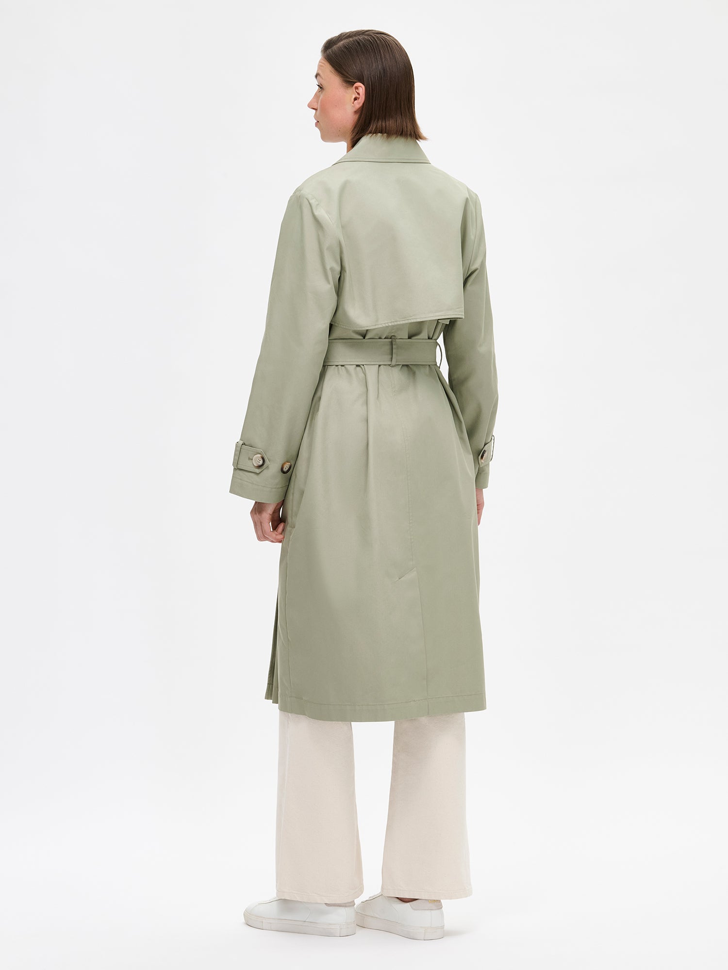 A timeless Light Green trench coat on a model with off white jeans pictured from the backside