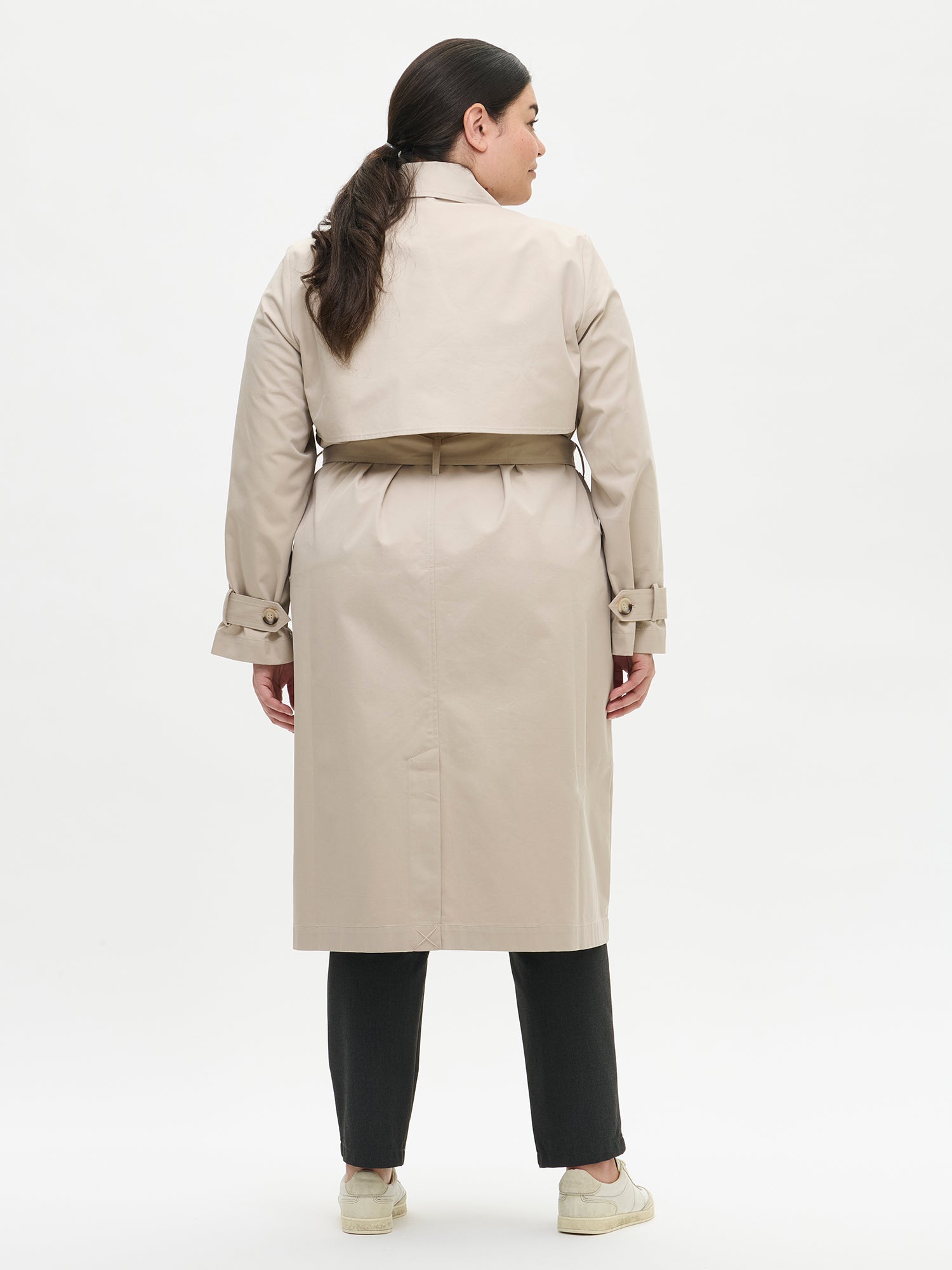 A timeless Beige trench coat on a model with black jeans pictured from the backside