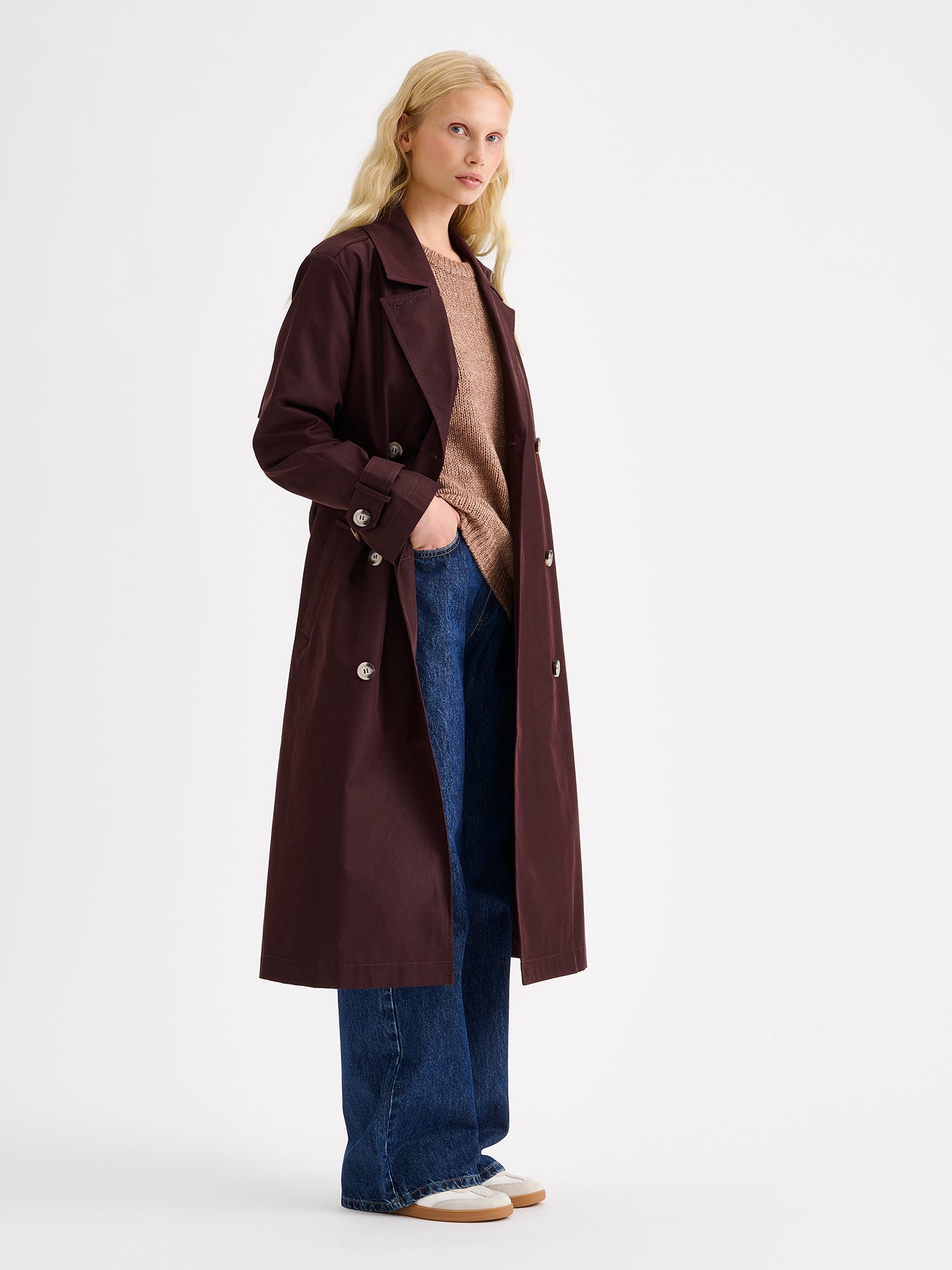 A timeless Brown trench coat on a blonde model with jeans and sneakers, the coat opened.