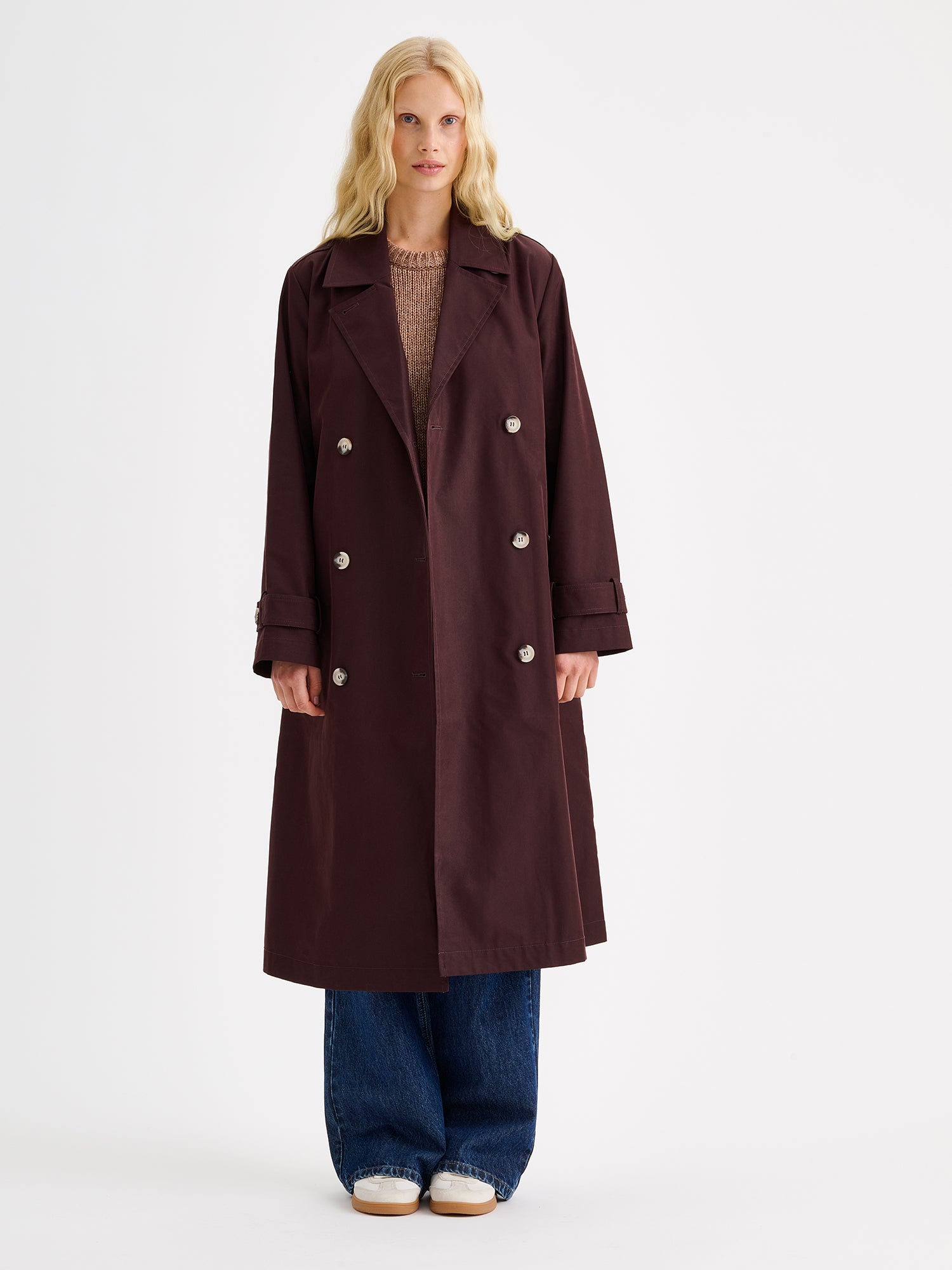 A timeless Brown trench coat on a blonde model with jeans and sneakers, the coat open.