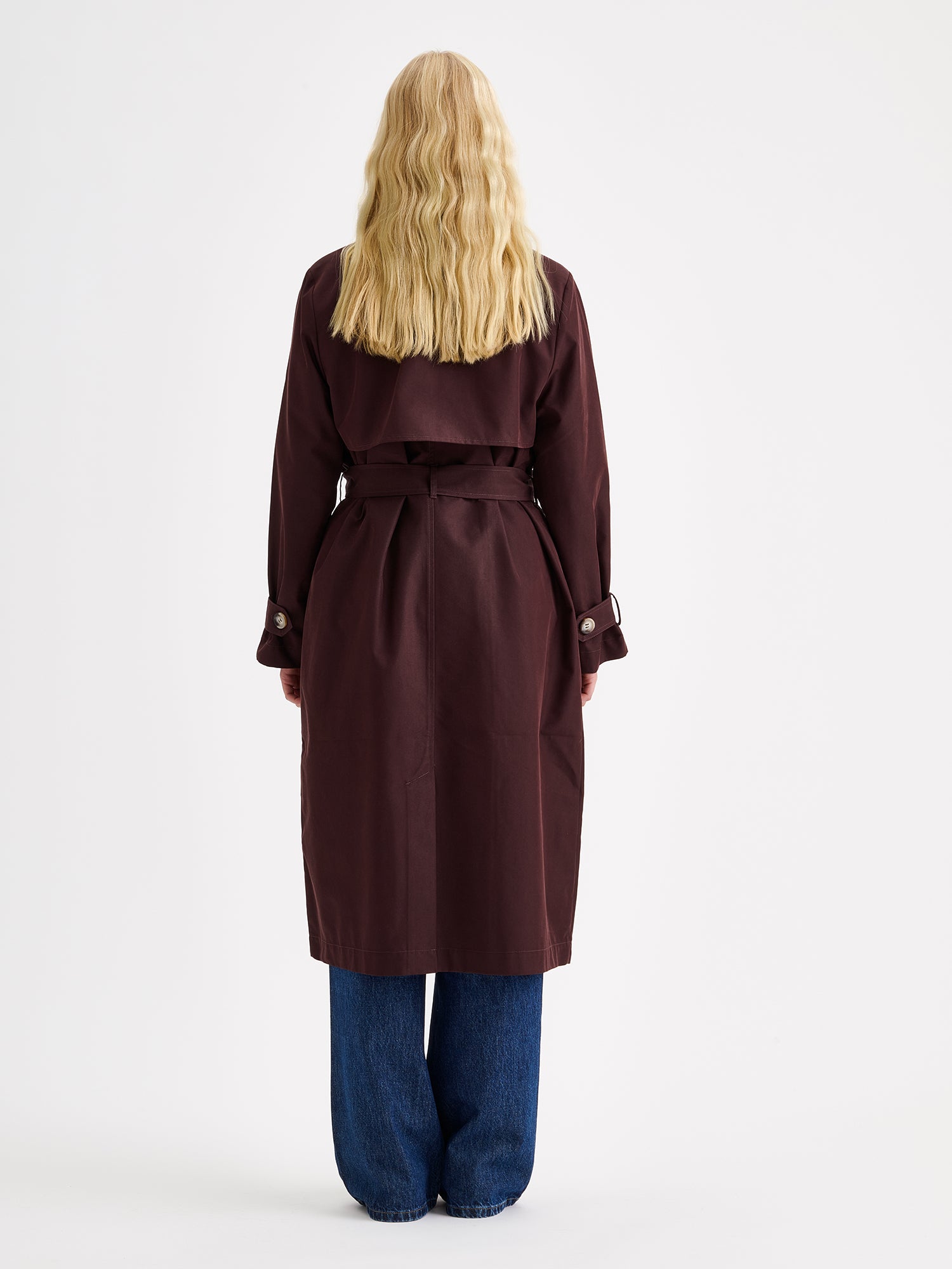 A timeless Brown trench coat on a blonde model with jeans and sneakers, pictured from the backside