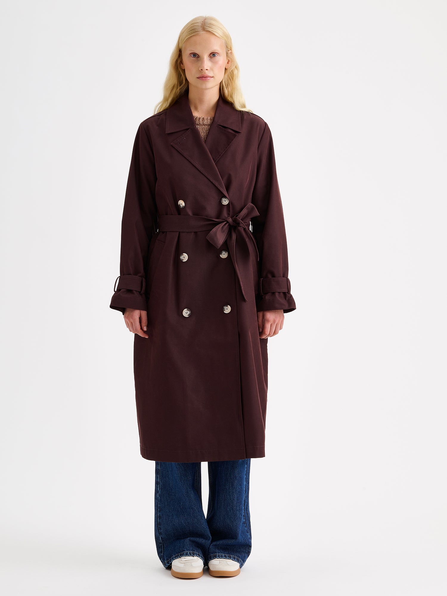 A timeless Brown trench coat on a blonde model with jeans and sneakers.