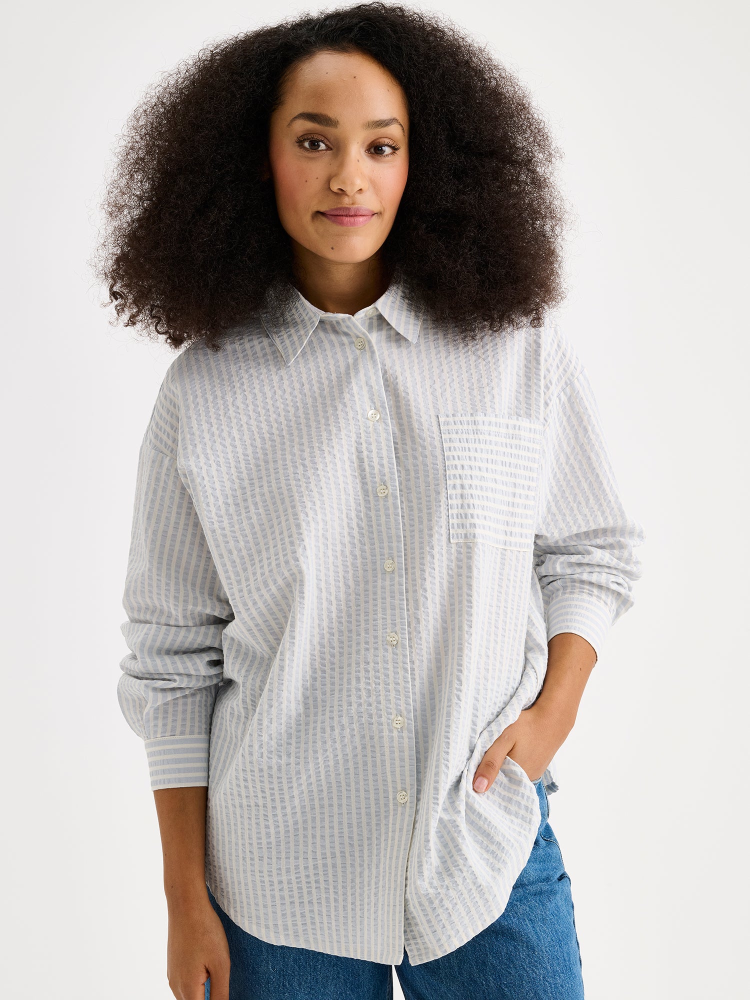 Nanso women's Marea Shirt in pale blue features a classic button-up shirt with a timeless and striped pattern. The shirt includes a collar, dropped shoulder line, chest pocket, and front buttons. On a model with jeans, close up.