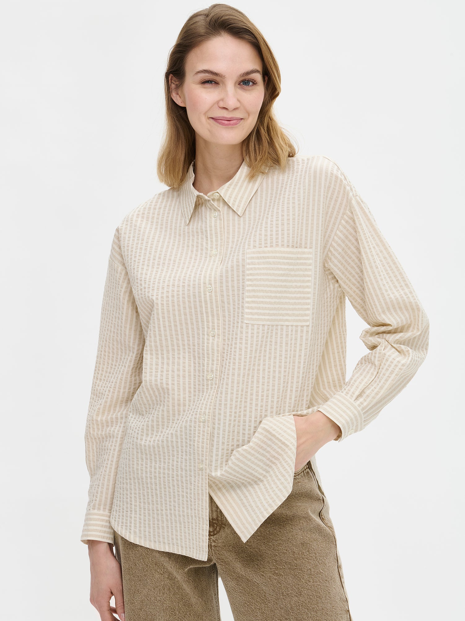 Nanso women's Marea Shirt in beige features a classic button-up shirt with a timeless and striped pattern. The shirt includes a collar, dropped shoulder line, chest pocket, and front buttons. On a model with jeans, close-up.
