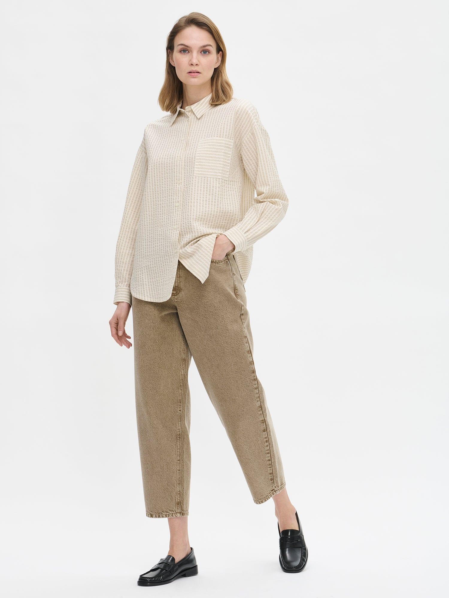 Nanso women's Marea Shirt in beige features a classic button-up shirt with a timeless and striped pattern. The shirt includes a collar, dropped shoulder line, chest pocket, and front buttons. On a model with jeans.