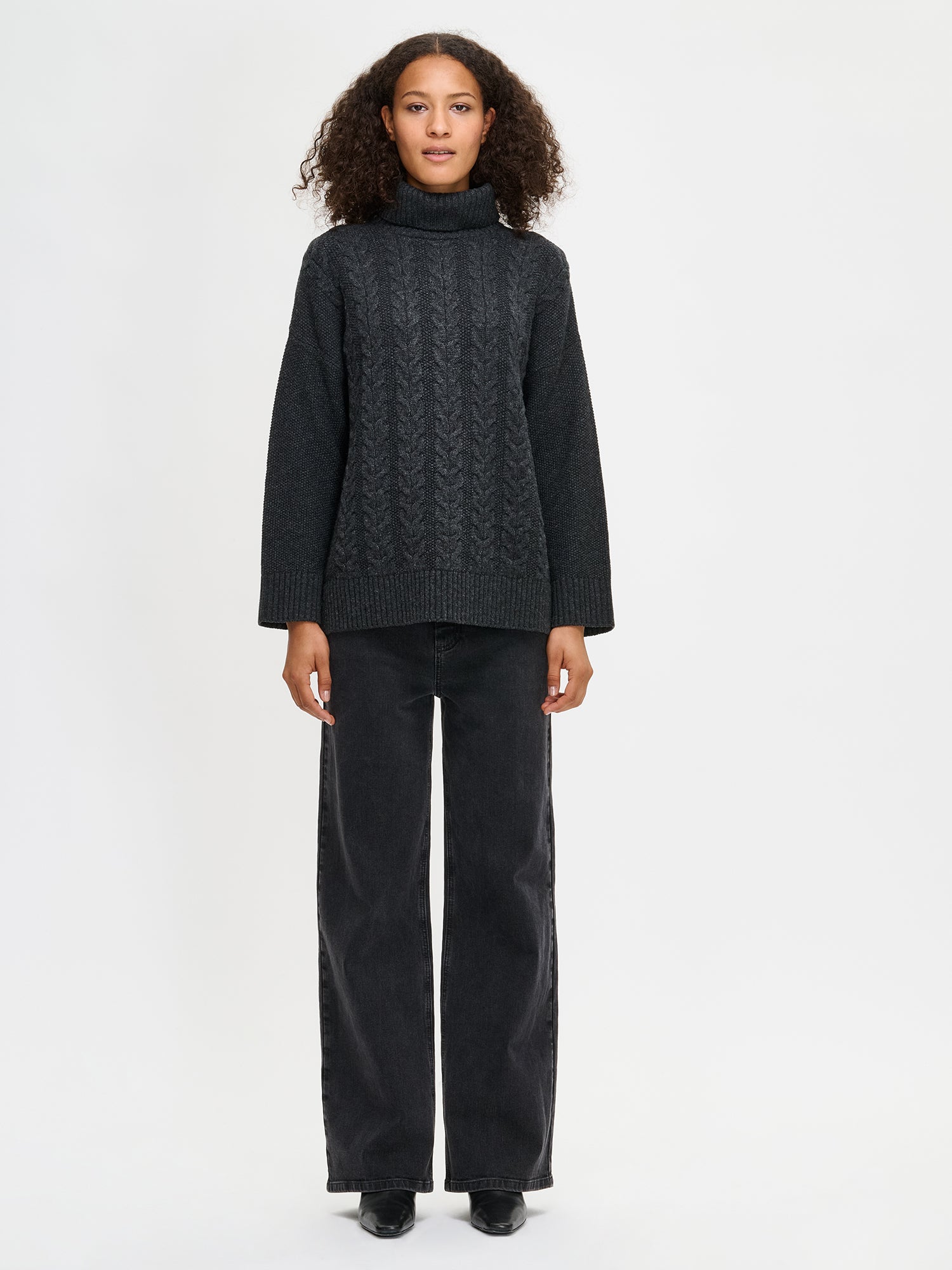 Woman wearing a dark gray cable knit sweater and black pants on a white background