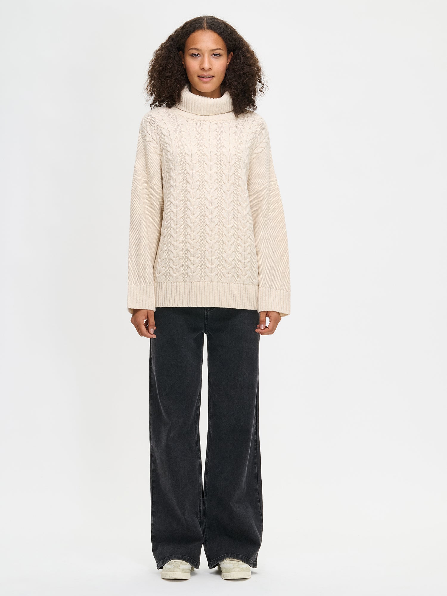 Woman wearing Nanso's a cream cable knit sweater and black pants on a white background