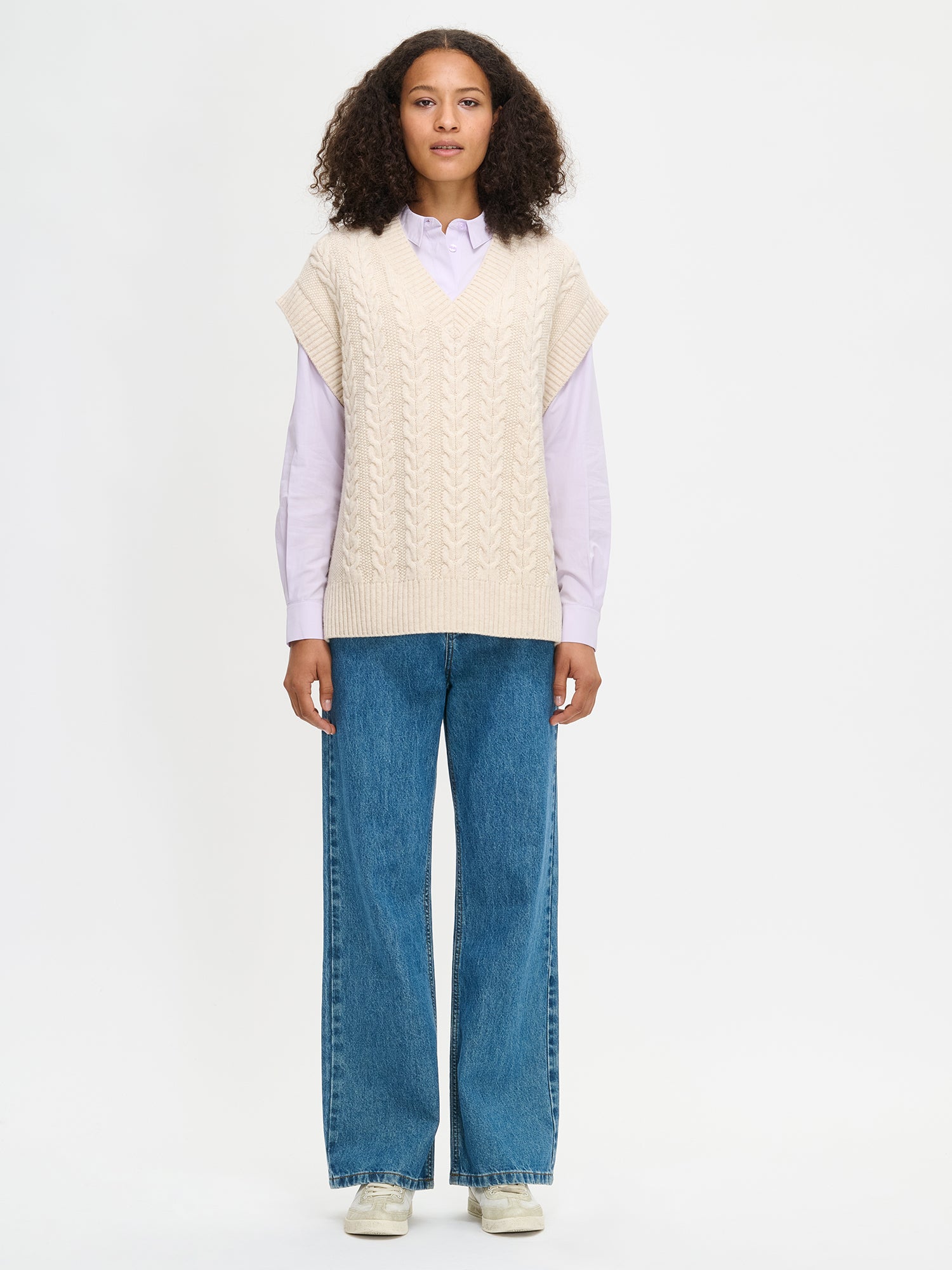 Woman wearing a cream vest over a light purple shirt with blue jeans on a white background