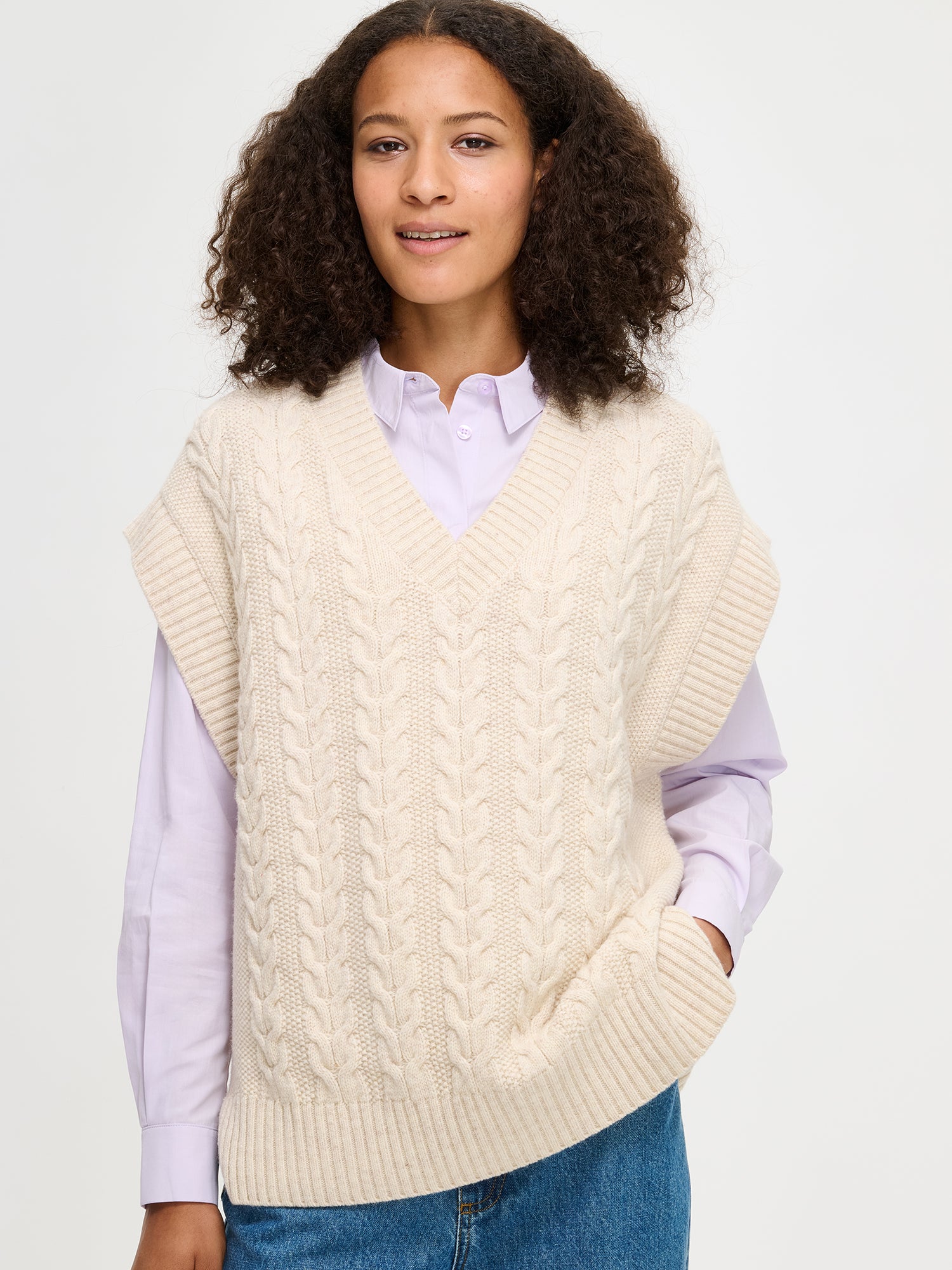 Woman wearing a cream cable knit vest over a light purple shirt on a white background
