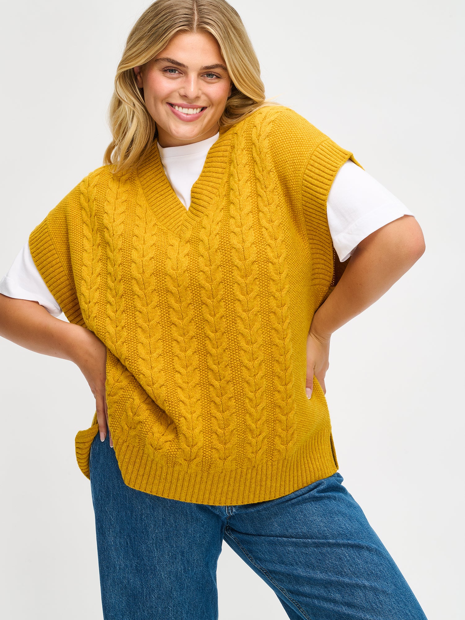 Woman wearing a yellow knitted vest over a white shirt and blue jeans on a light gray background