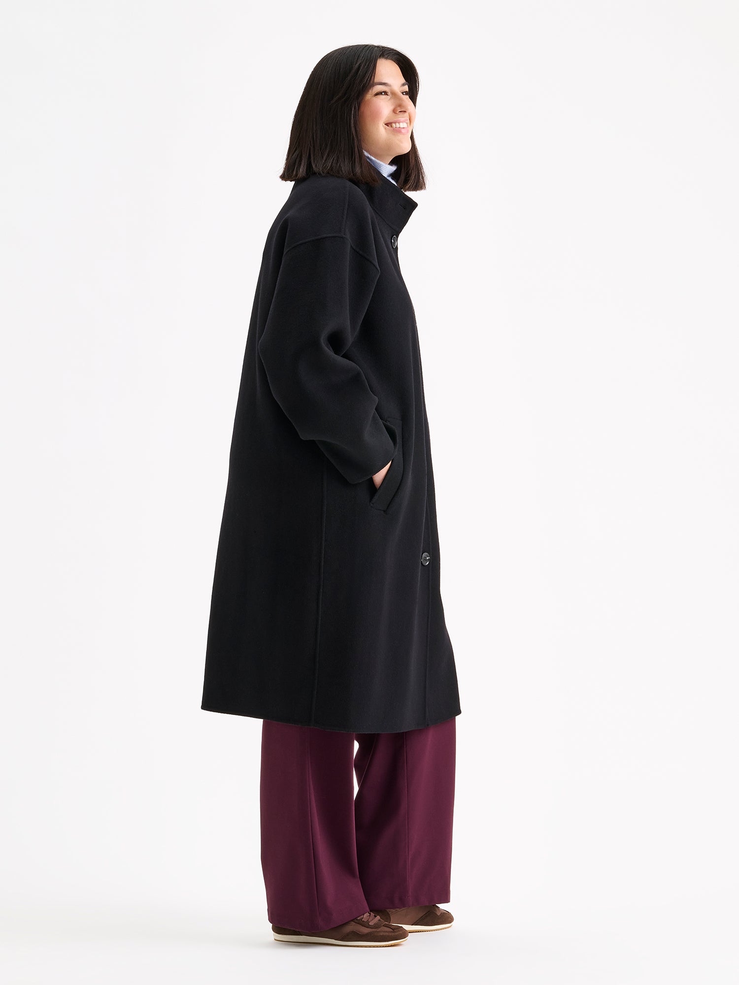 Nanso women's long black Elle wool coat on a brunette model with a light blue scarf and winered jeans, side angle.