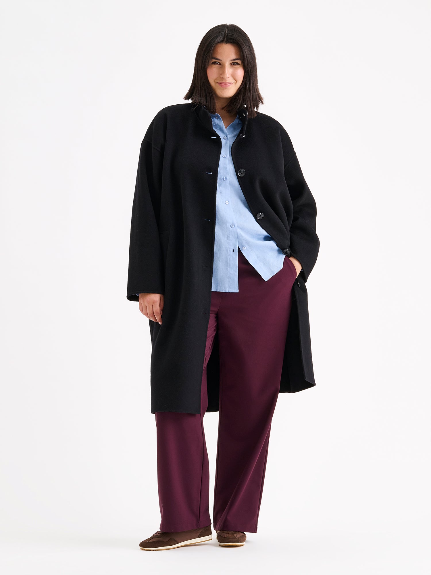 Nanso women's long black Elle wool coat on a brunette model with a light blue shirt and winered jeans. | XL - 178cm