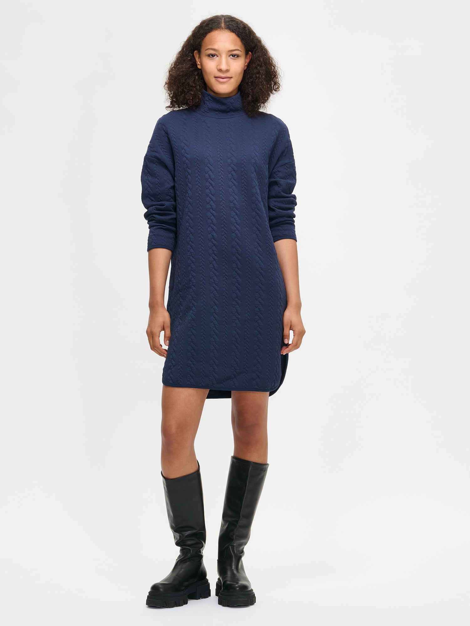 Woman wearing a dark blue dress with a turtleneck and black boots on a white background
