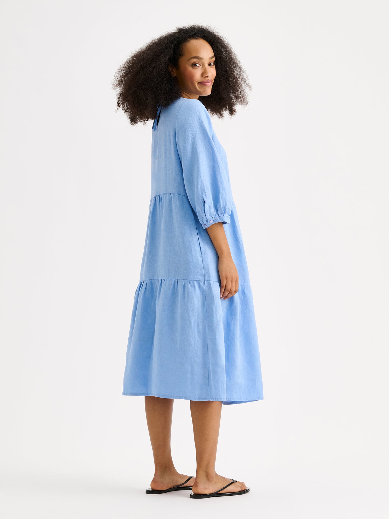 Nanso women's Saralia Linen Dress in blue with voluminous frill layers on a model with flip flops, photographed from side angle.