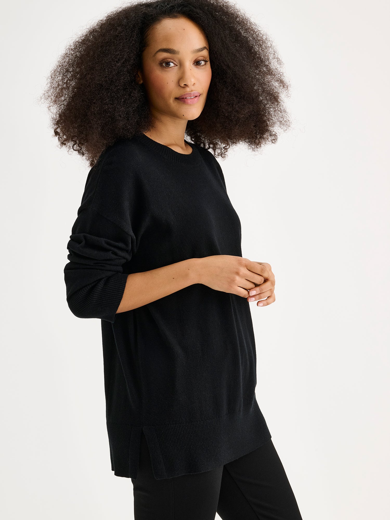 The VILLIS Long Knit Sweater in black features a timeless boxy silhouette and a classic crew neckline, dropped shoulders, and full-length sleeves and ribbed trim at the hem and cuffs. On a model pictured from the side.