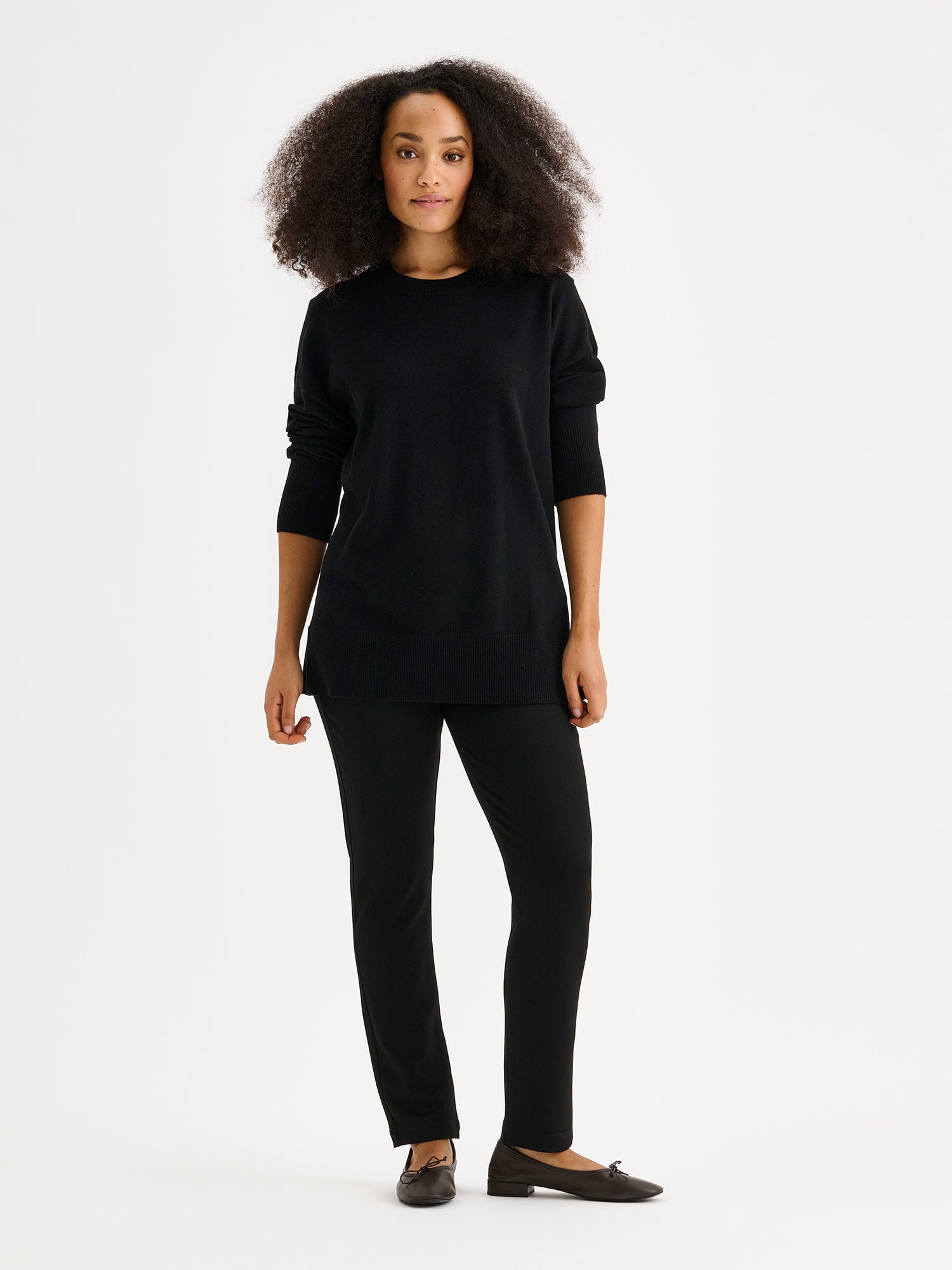 The VILLIS Long Knit Sweater in black features a timeless boxy silhouette and a classic crew neckline, dropped shoulders, and full-length sleeves and ribbed trim at the hem and cuffs. On a model.