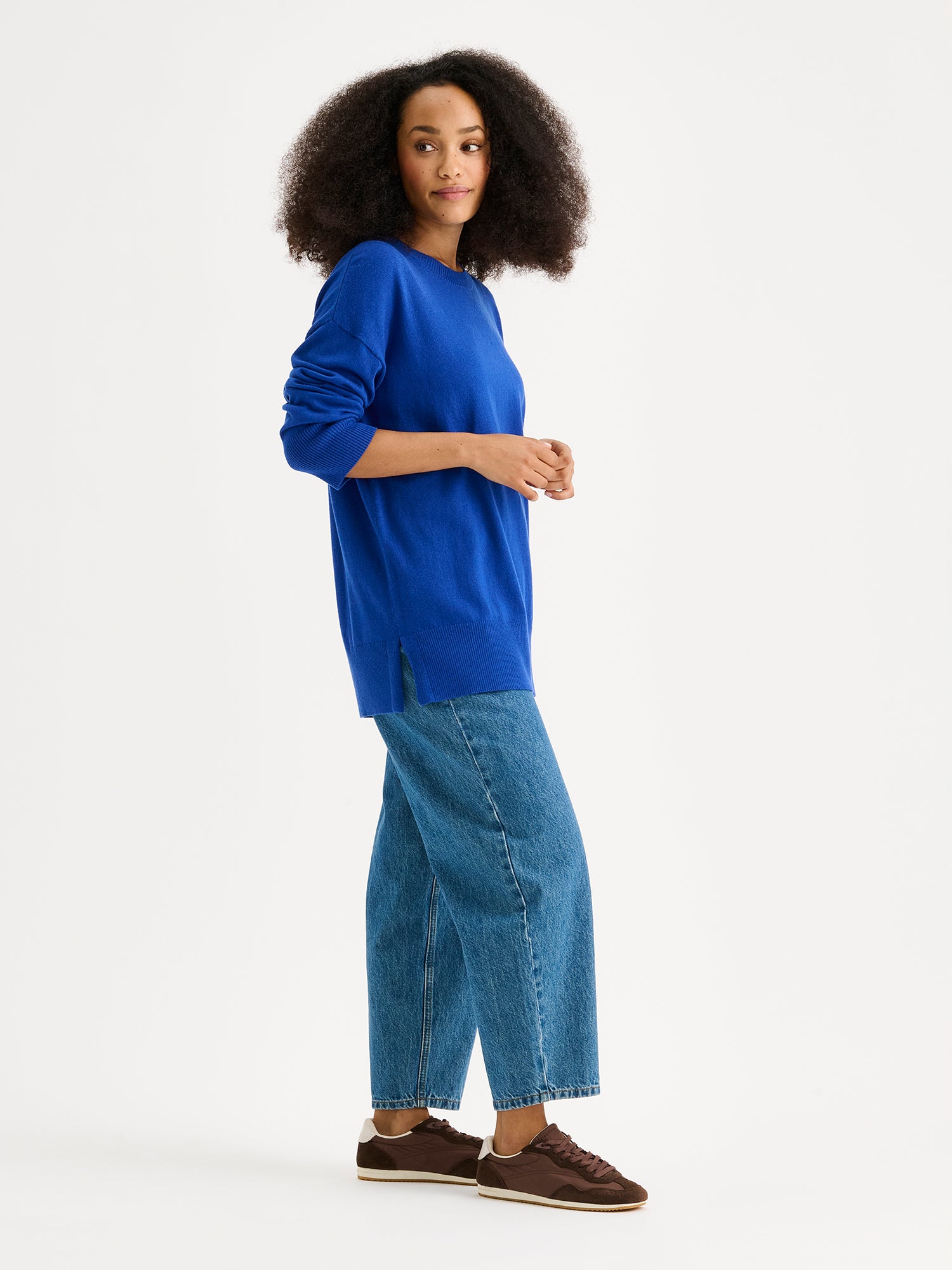 The VILLIS Long Knit Sweater in blue features a timeless boxy silhouette and a classic crew neckline, dropped shoulders, and full-length sleeves and ribbed trim at the hem and cuffs. On a model with blue jeans from the side.