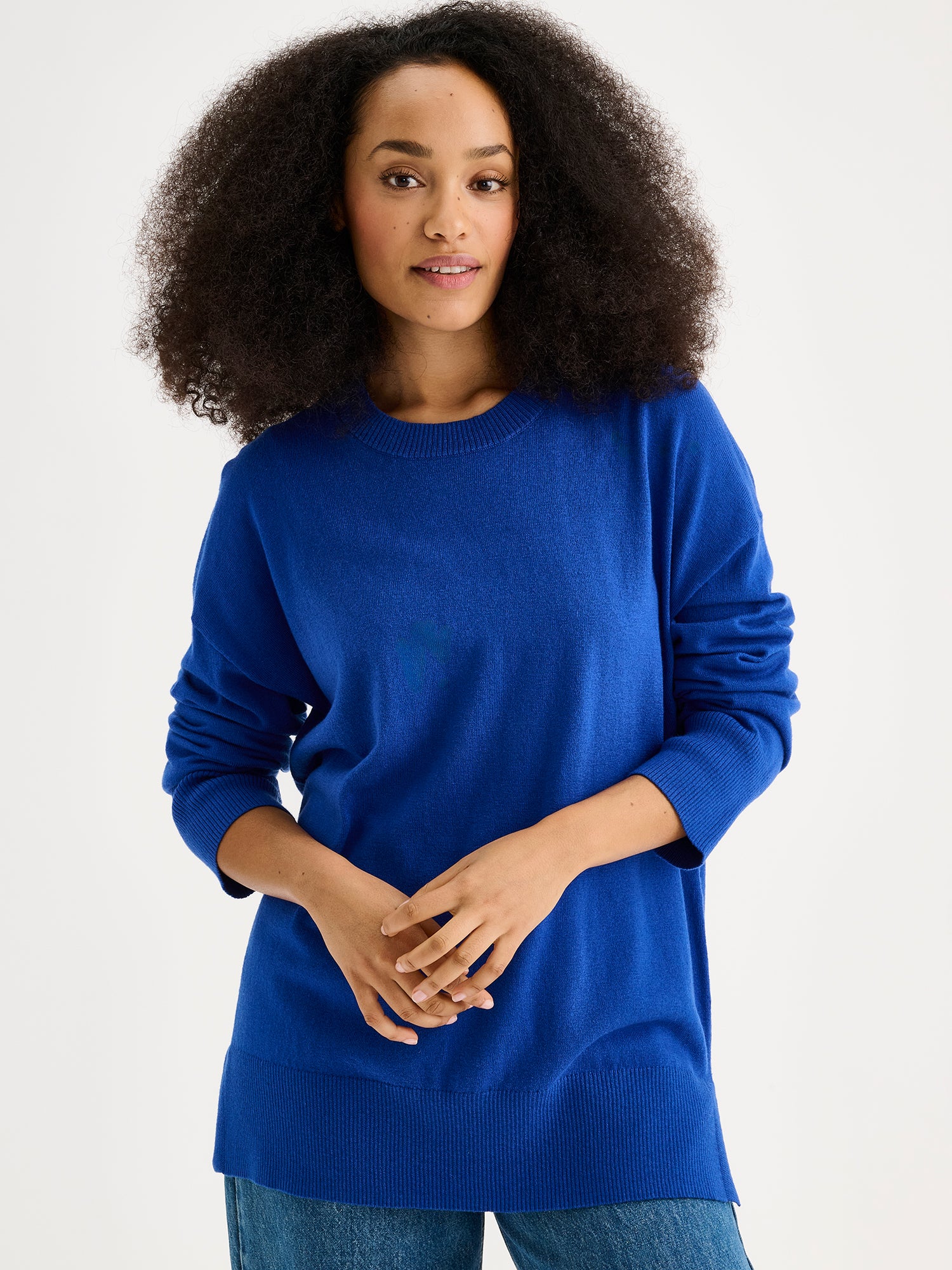 The VILLIS Long Knit Sweater in blue features a timeless boxy silhouette and a classic crew neckline, dropped shoulders, and full-length sleeves and ribbed trim at the hem and cuffs. On a model with blue jeans, close up.