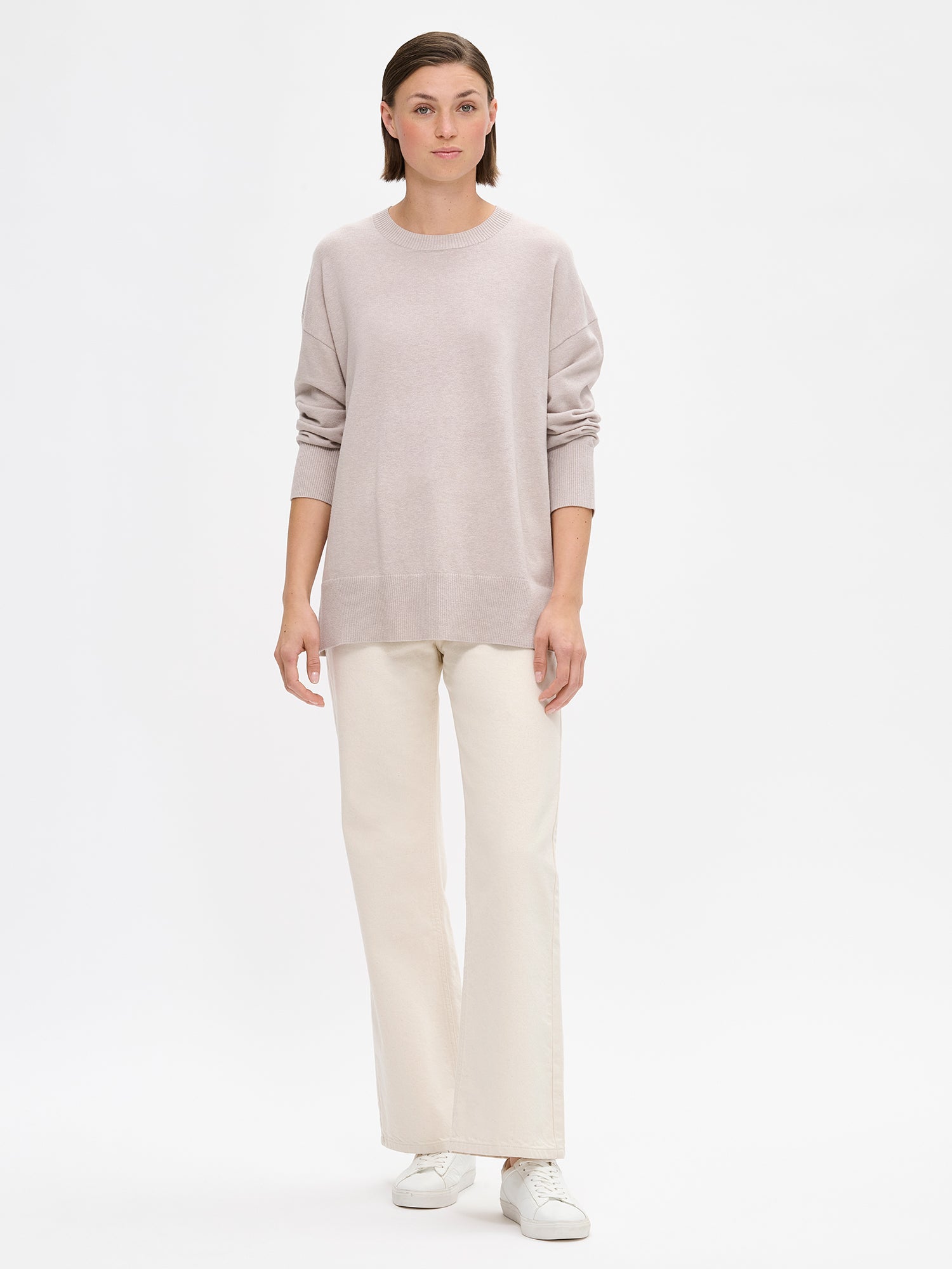 The VILLIS Long Knit Sweater in greige features a timeless boxy silhouette and a classic crew neckline, dropped shoulders, and full-length sleeves and ribbed trim at the hem and cuffs. On a model with off white jeans and sneakers.