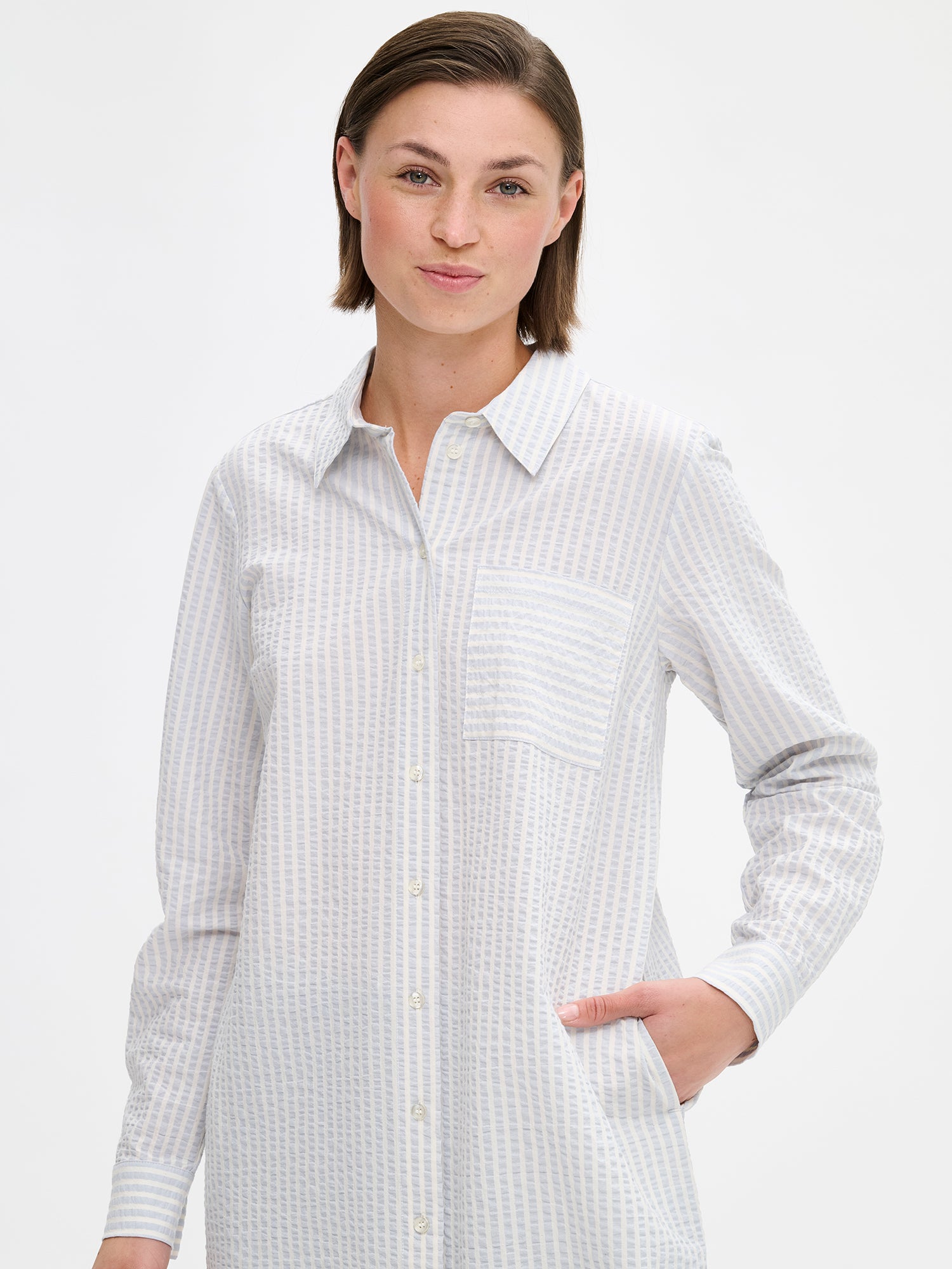 Nanso women's MAREA shirt dress in pale blue features a classic collar, front button closure, rounded hemline. A chest pocket and side pockets add everyday functionality. On a model, close-up.