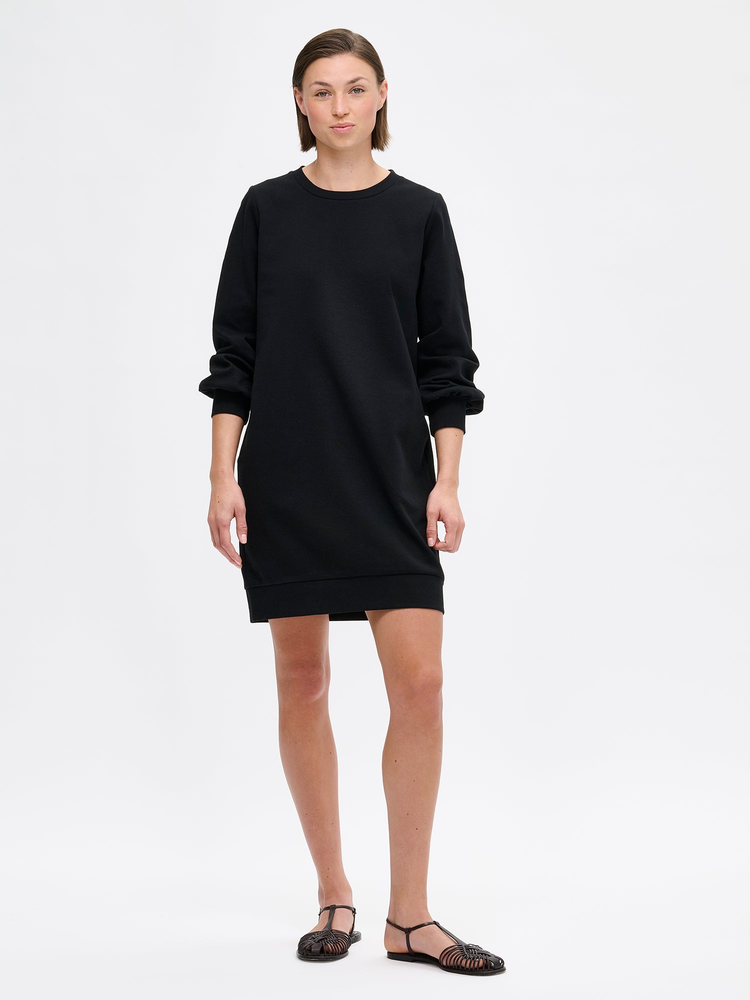Nanso women's black Leija-dress. This straight-cut dress features a round neckline, side pockets, and full-length sleeves that gather beautifully at the cuffs. On a model with sandals.