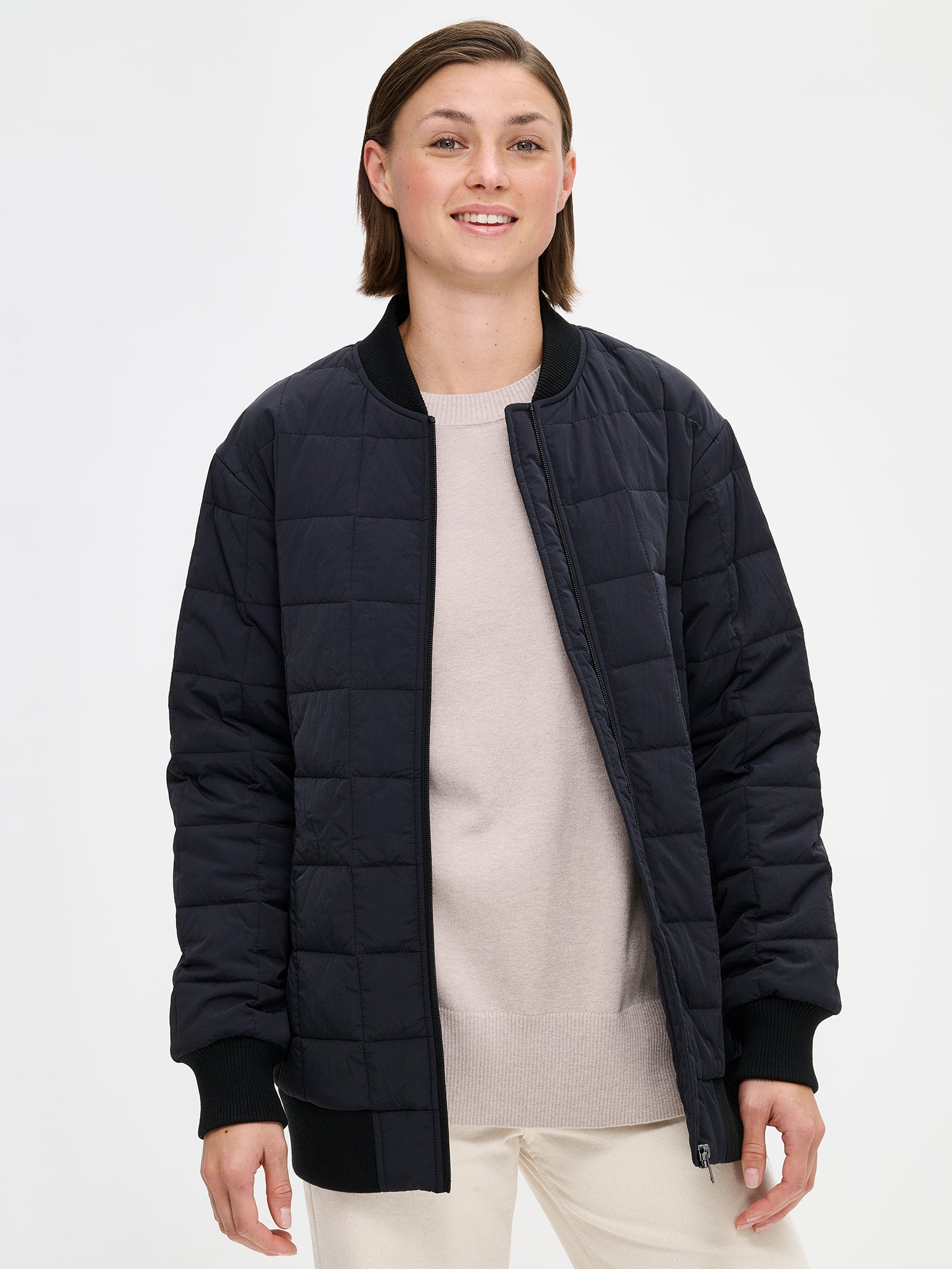 Nanso MERJE Quilted Bomber Jacket in Black features a lightweight Sorona -insulation, ribbed knit detailing at the neckline, cuffs, and hem. Dropped shoulders create a relaxed, contemporary silhouette. Photographed on a model with jeans on, jacket opened close-up.