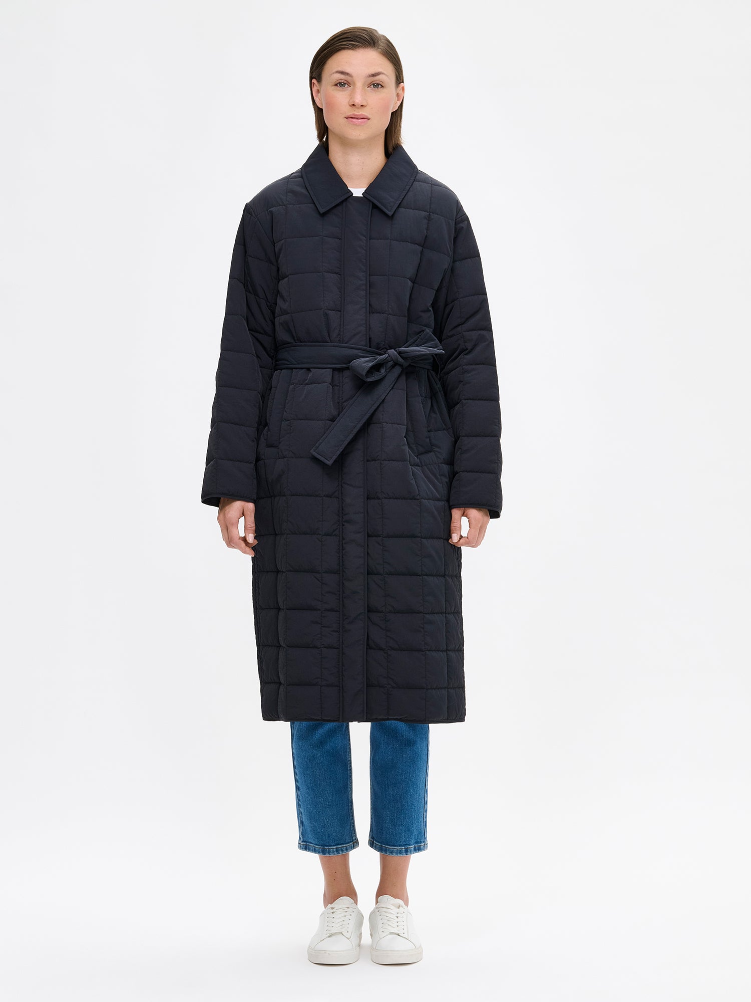 Nanso Marje Quilted Coat features classic collar styling and convenient snap closures at the front, a belt for a personalized fit, and practical front patch pockets. On a model with jeans.