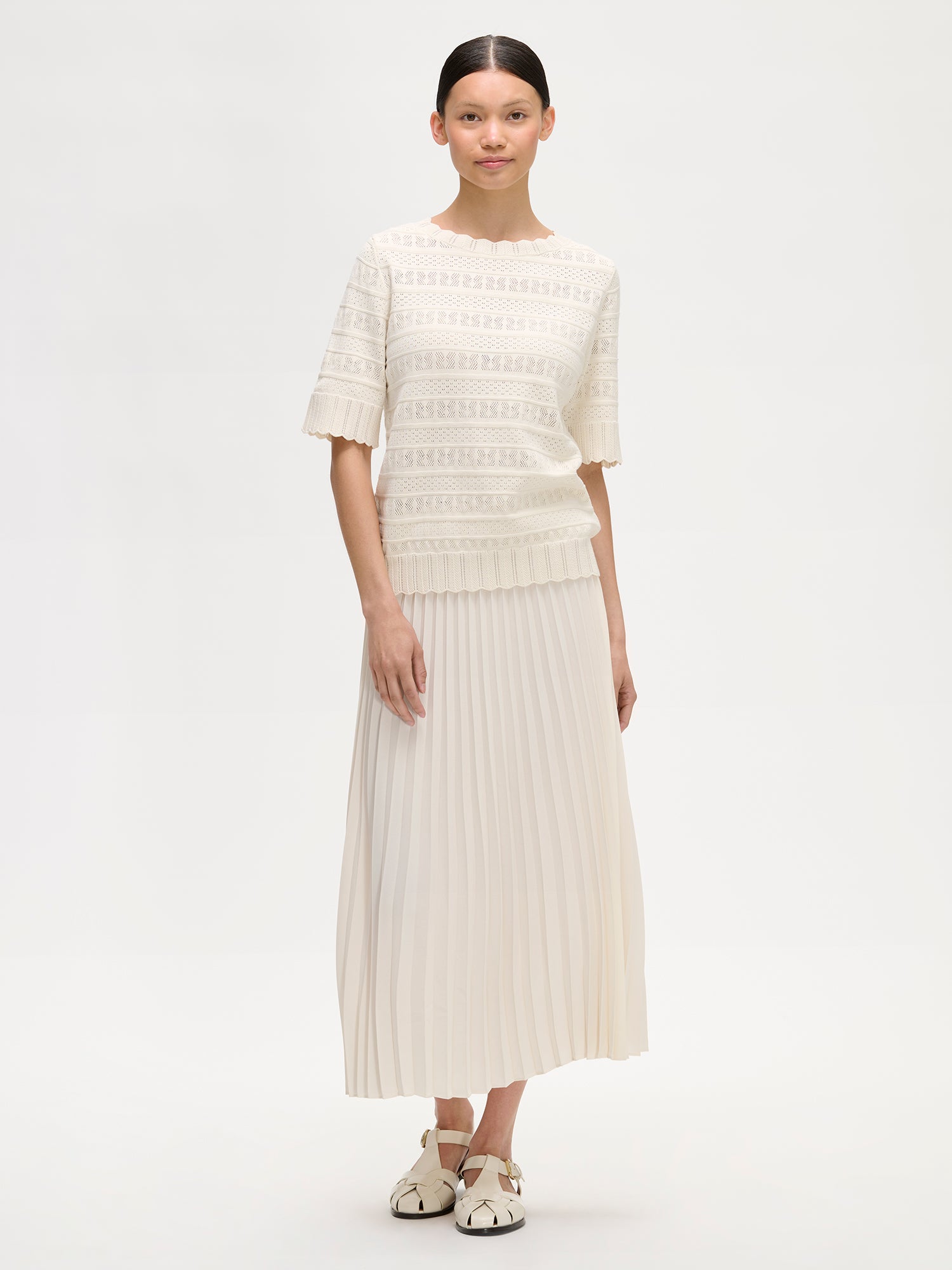 Nanso timeless white KORU knit sweater featuring delicate lace knit that elevates any wardrobe on a model with white pleated skirt