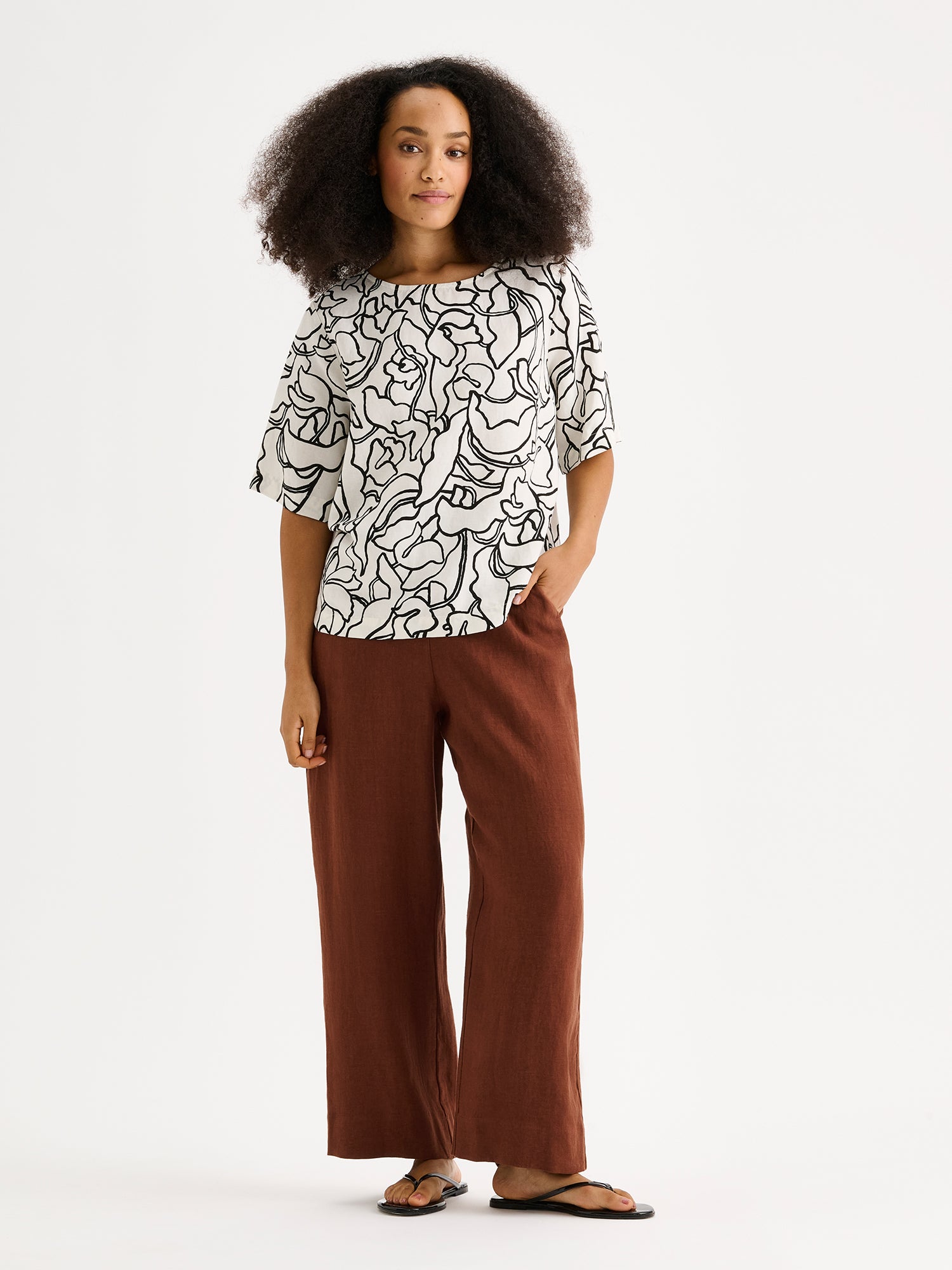 Nanso women's stylish and wonderfully relaxed IIRIS blouse featuring half-length sleeves and an elegant black and white floral pattern on a model with brown linen trousers.