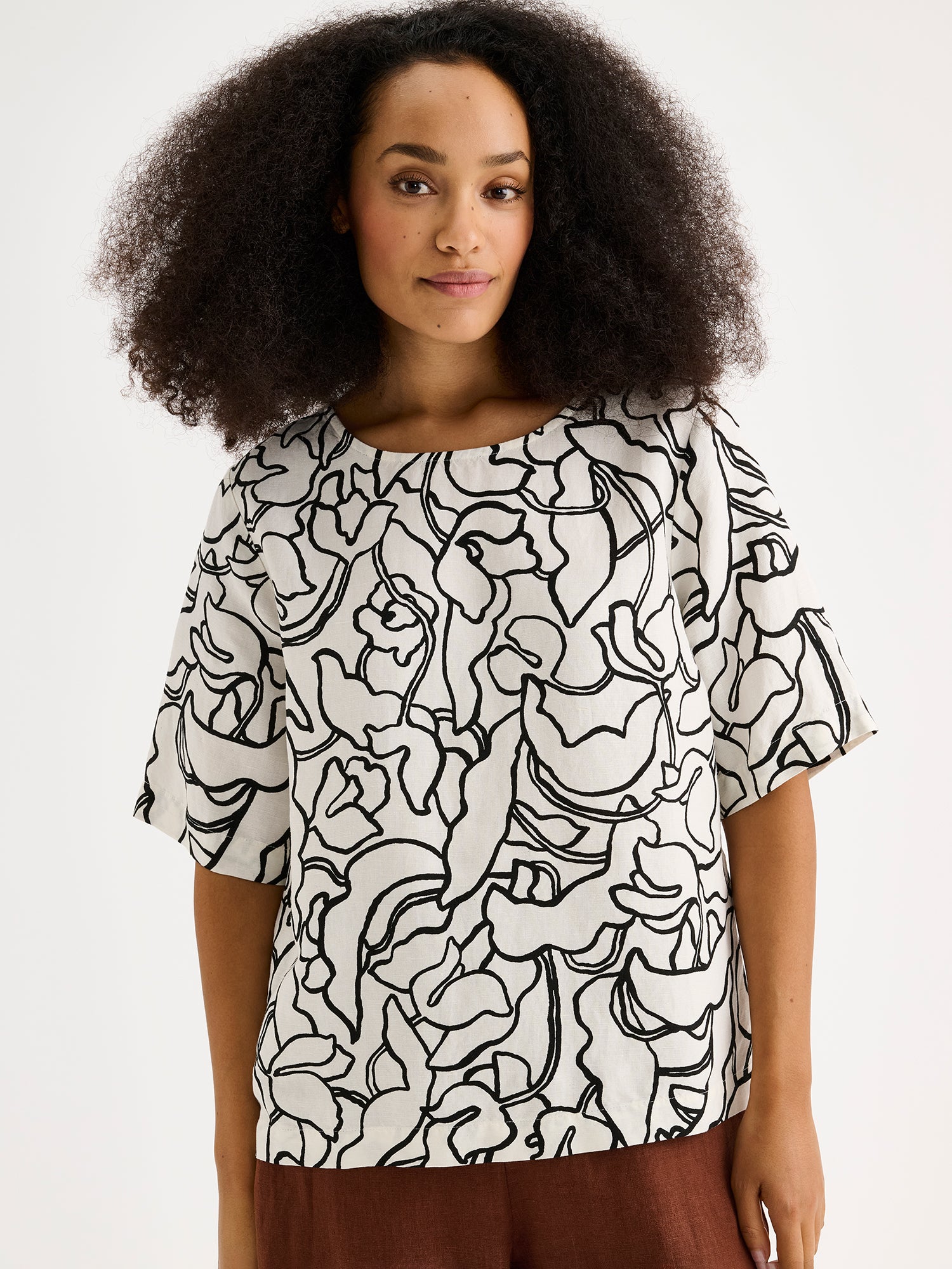 Nanso women's stylish and wonderfully relaxed IIRIS blouse featuring half-length sleeves and an elegant black and white floral pattern on a model with brown linen trousers, close-up.