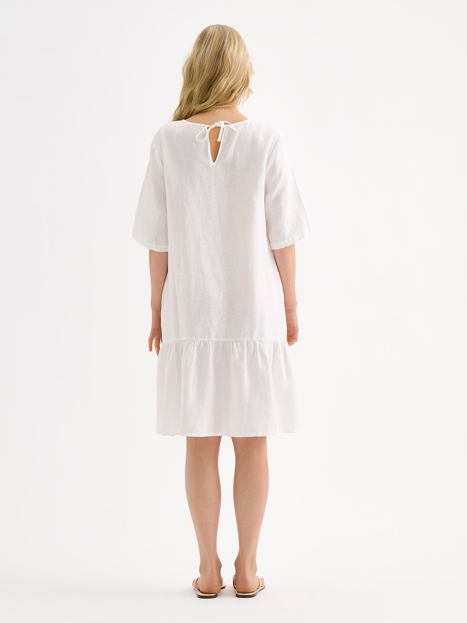 Nanso women's white relaxed Liisu linen dress with half-length sleeves features a generously ruffled hem on a model with sandals, pictured from the backside