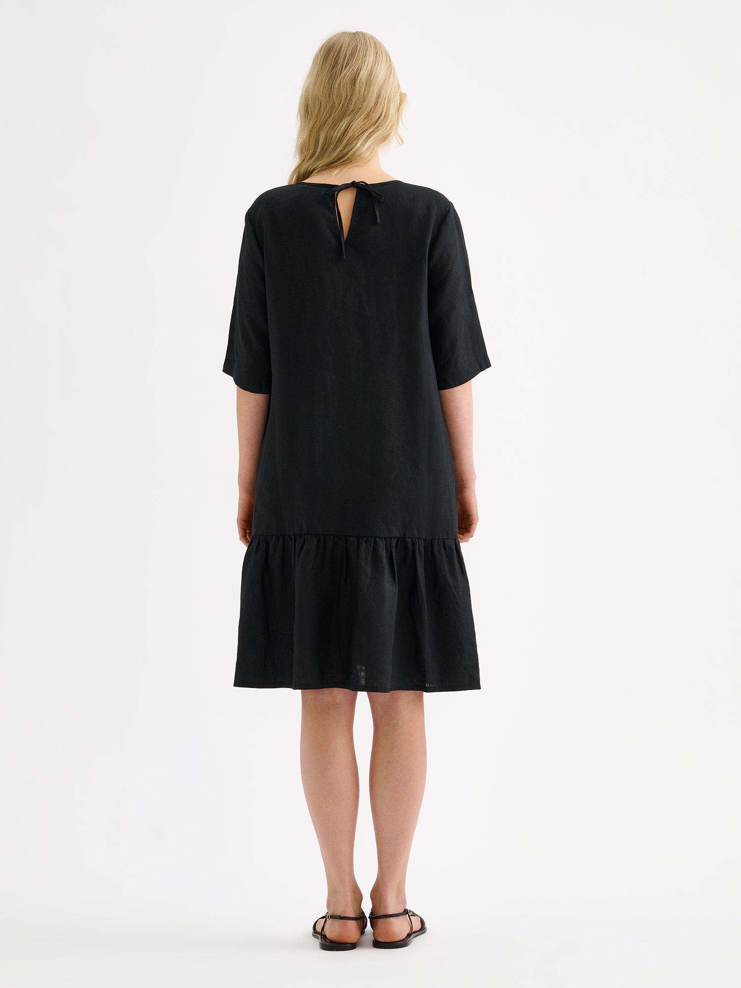 Nanso women's black relaxed Liisu linen dress with half-length sleeves features a generously ruffled hem, on a model pictured from the backside