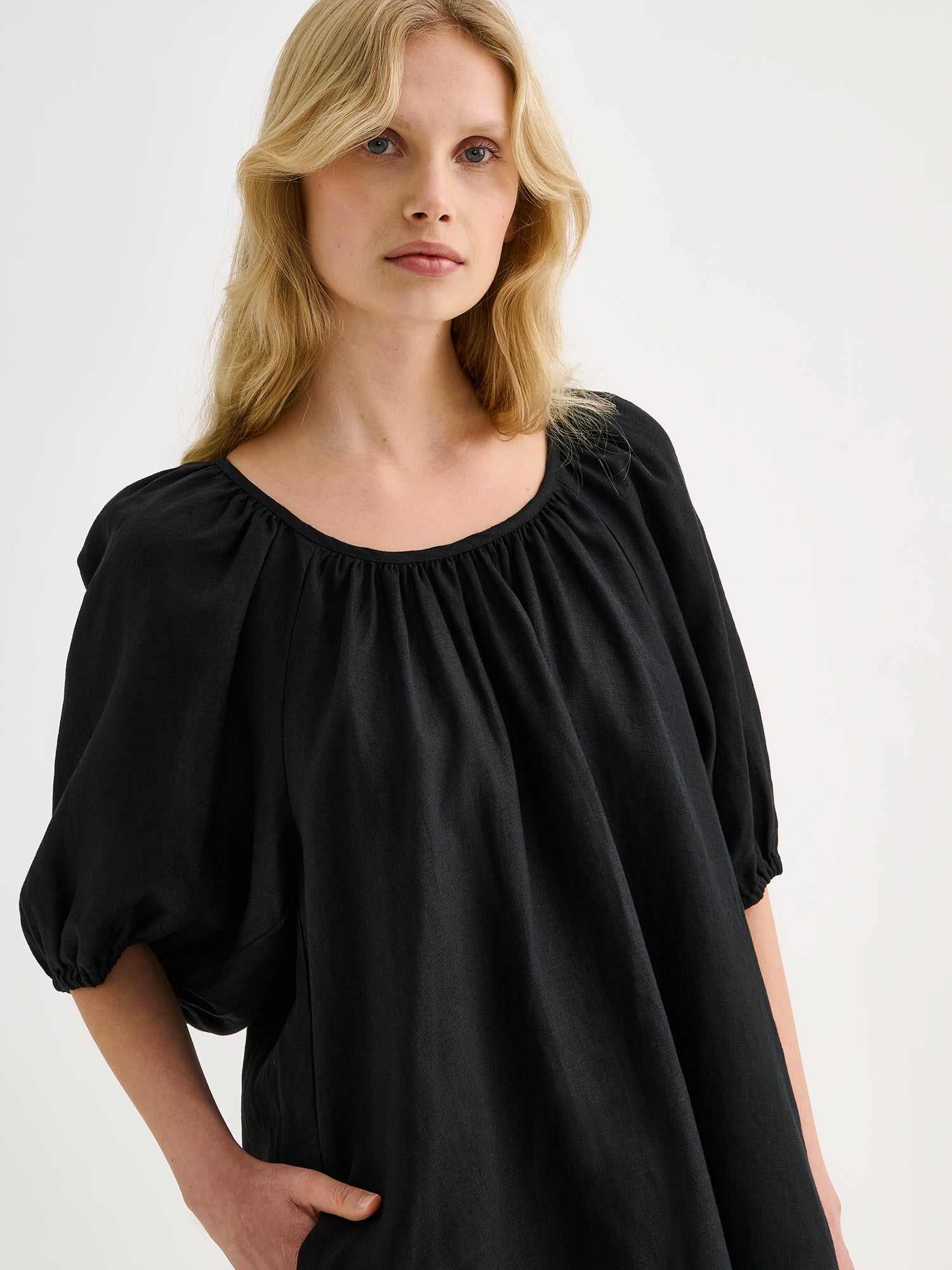 Nanso women’s beautifully relaxed black Donna linen dress. This airy short dress features a softly gathered open back neckline, finished with a tie-back bow detail. Pictured on a model, close-up.