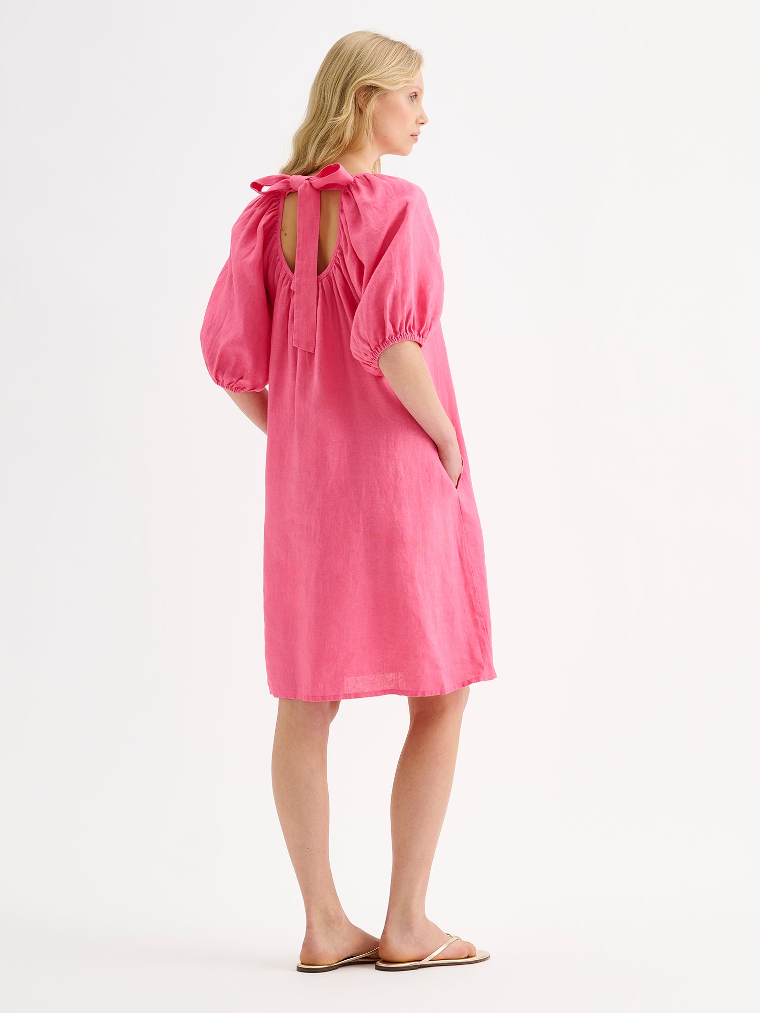 Nanso women’s beautifully relaxed pink Donna linen dress. This airy short dress features a softly gathered open back neckline, finished with a tie-back bow detail. Pictured on a model with sandals from the backside