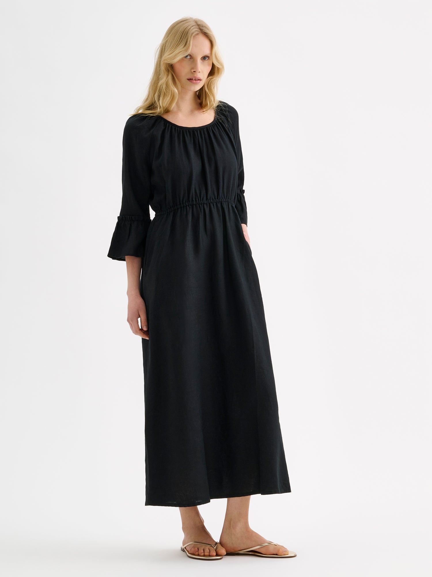 Nanso women's black  Donna midi linen dress combines effortless elegance with feminine details. This airy midi-length style features an open, softly gathered back neckline with a striking self-tie bow. Pictured on a blonde model with golden sandals.