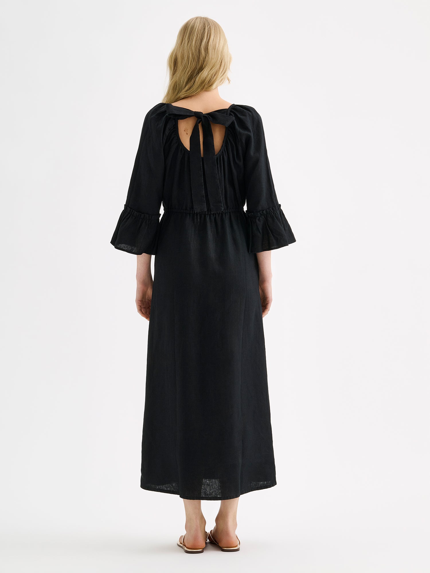 Nanso women's black  Donna midi linen dress combines effortless elegance with feminine details. This airy midi-length style features an open, softly gathered back neckline with a striking self-tie bow. Pictured on a blonde model with golden sandals, from the backside.