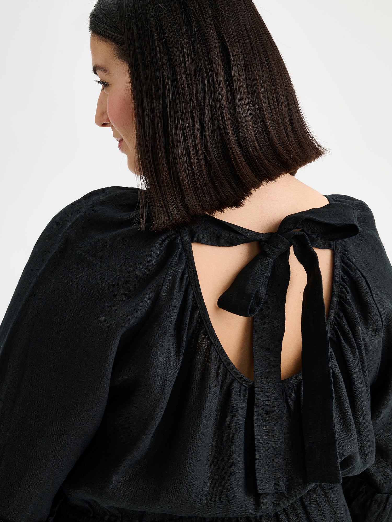 Nanso women's black  Donna midi linen dress combines effortless elegance with feminine details. This airy midi-length style features an open, softly gathered back neckline with a striking self-tie bow. Pictured on a model, close-up from the backside.