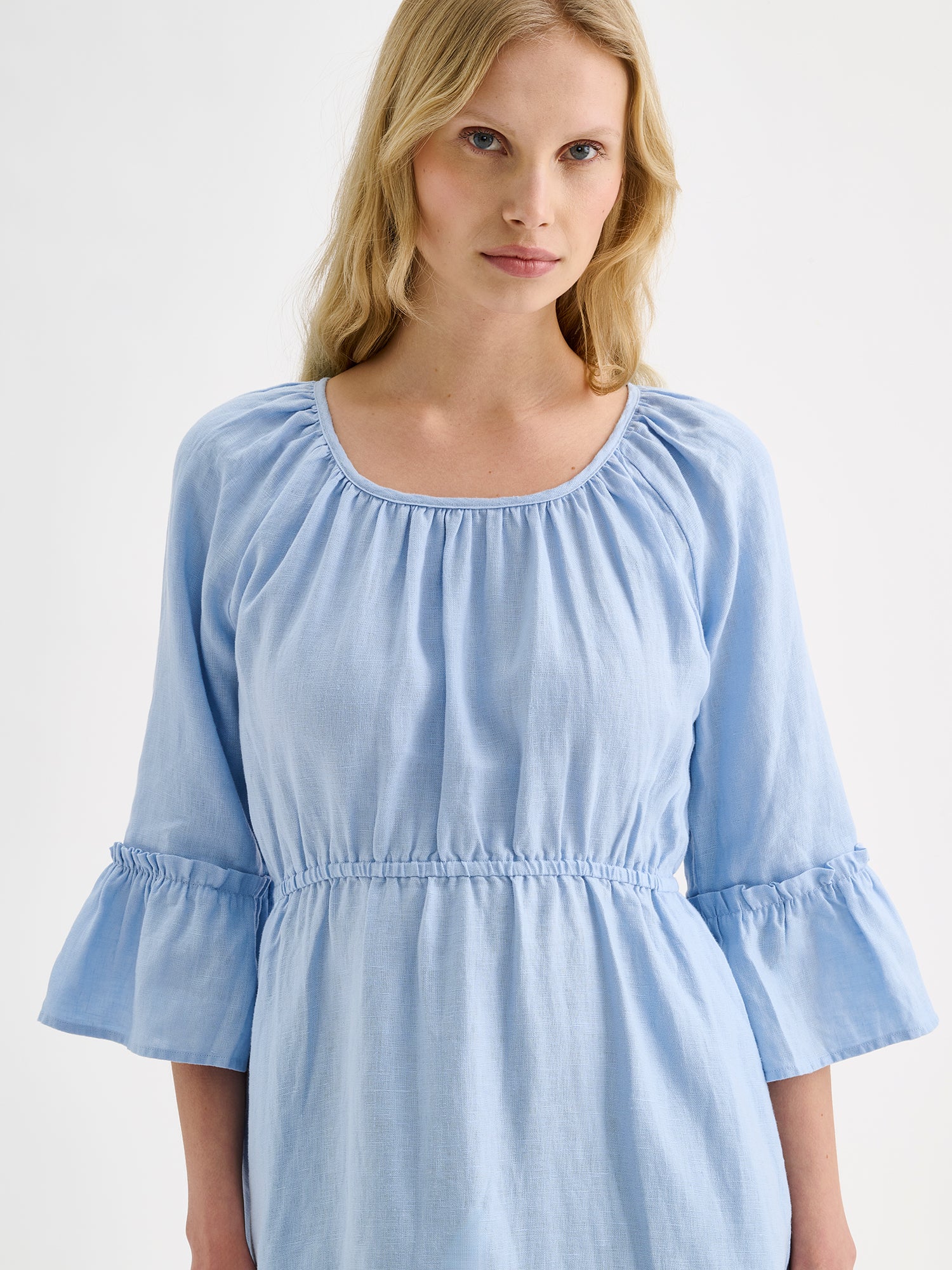 Nanso women's softly light blue Donna midi linen dress combines effortless elegance with feminine details. This airy midi-length style features an open, softly gathered back neckline with a striking self-tie bow. Pictured on a blonde model, close-up.