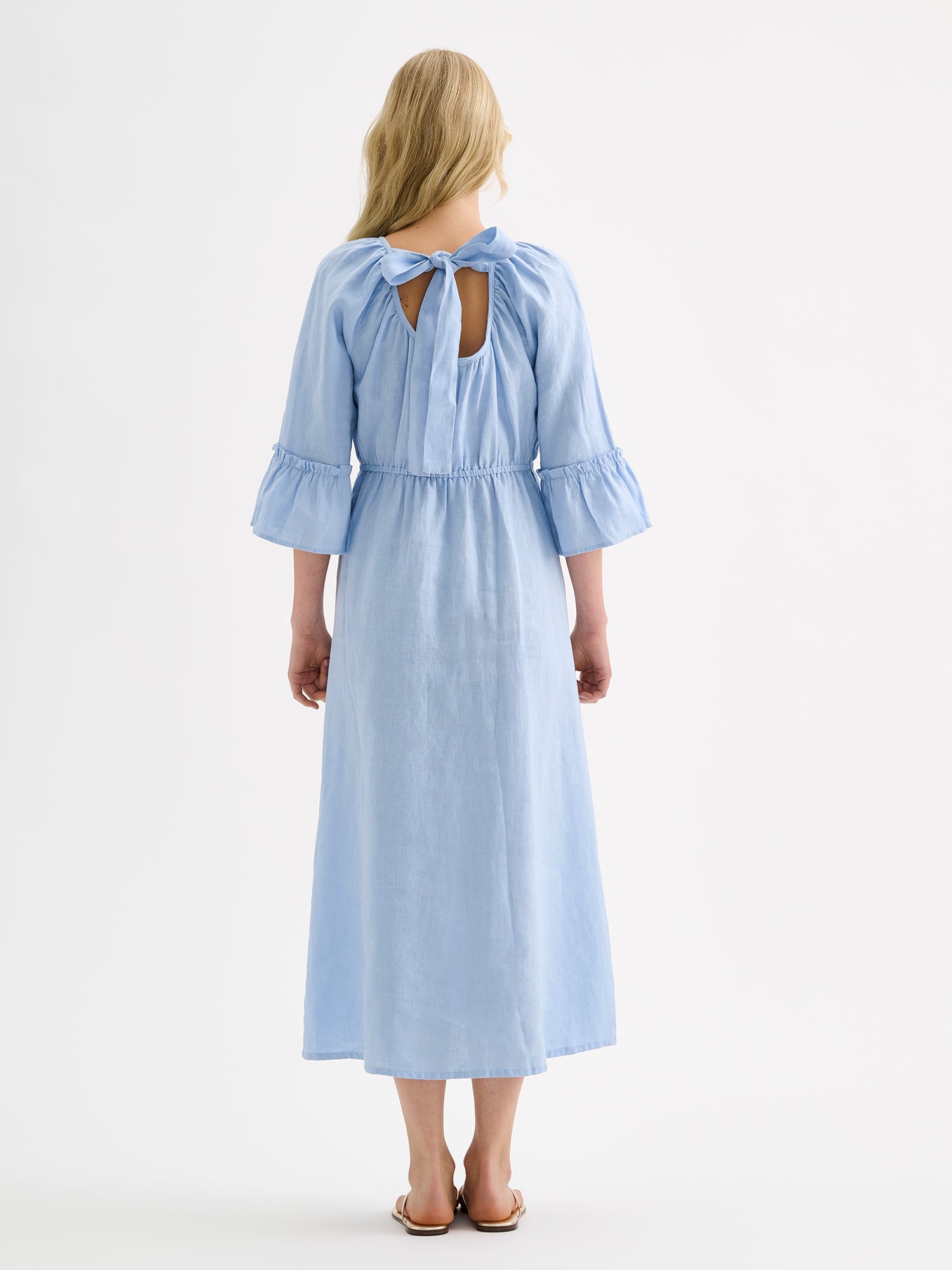 Nanso women's softly light blue Donna midi linen dress combines effortless elegance with feminine details. This airy midi-length style features an open, softly gathered back neckline with a striking self-tie bow. Pictured on a blonde model with golden sandals, from the backside.
