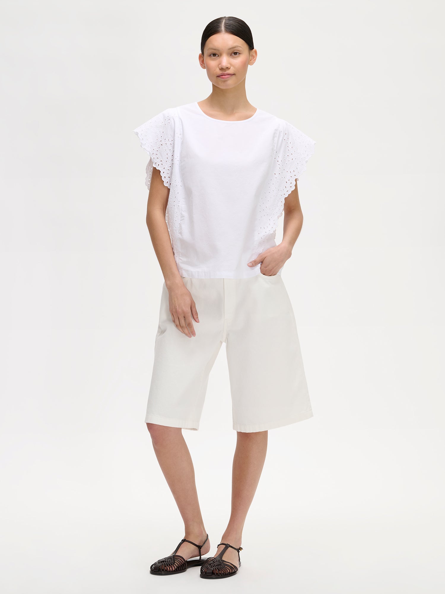 Nanso women's white Sevilla blouse for women combines an airy silhouette with delicate lace details. On a model with white shorts and sandals.