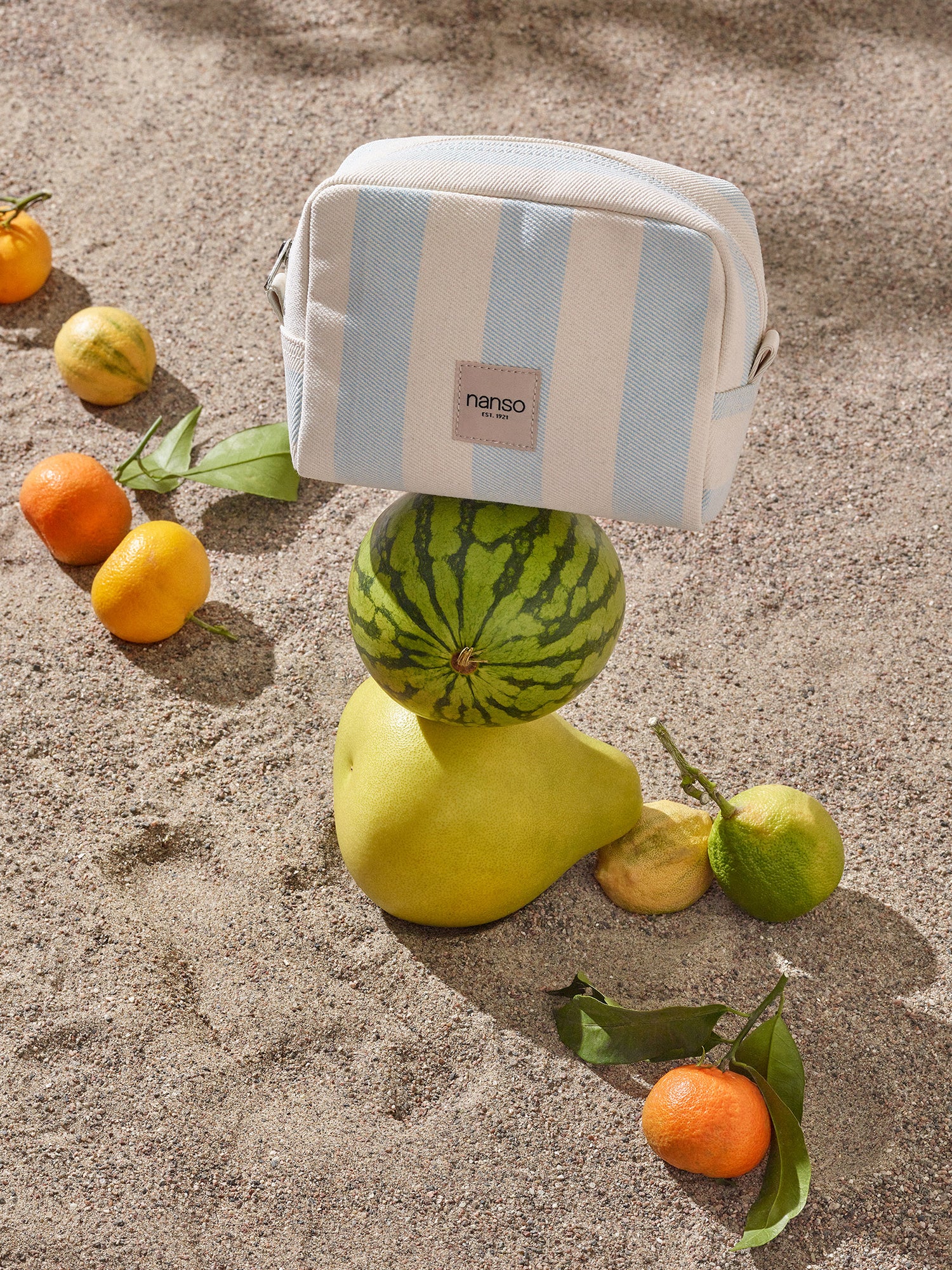 The AMALFI MINI cosmetics bag in Light Blue in a fresh product picture on sand with fruits.
