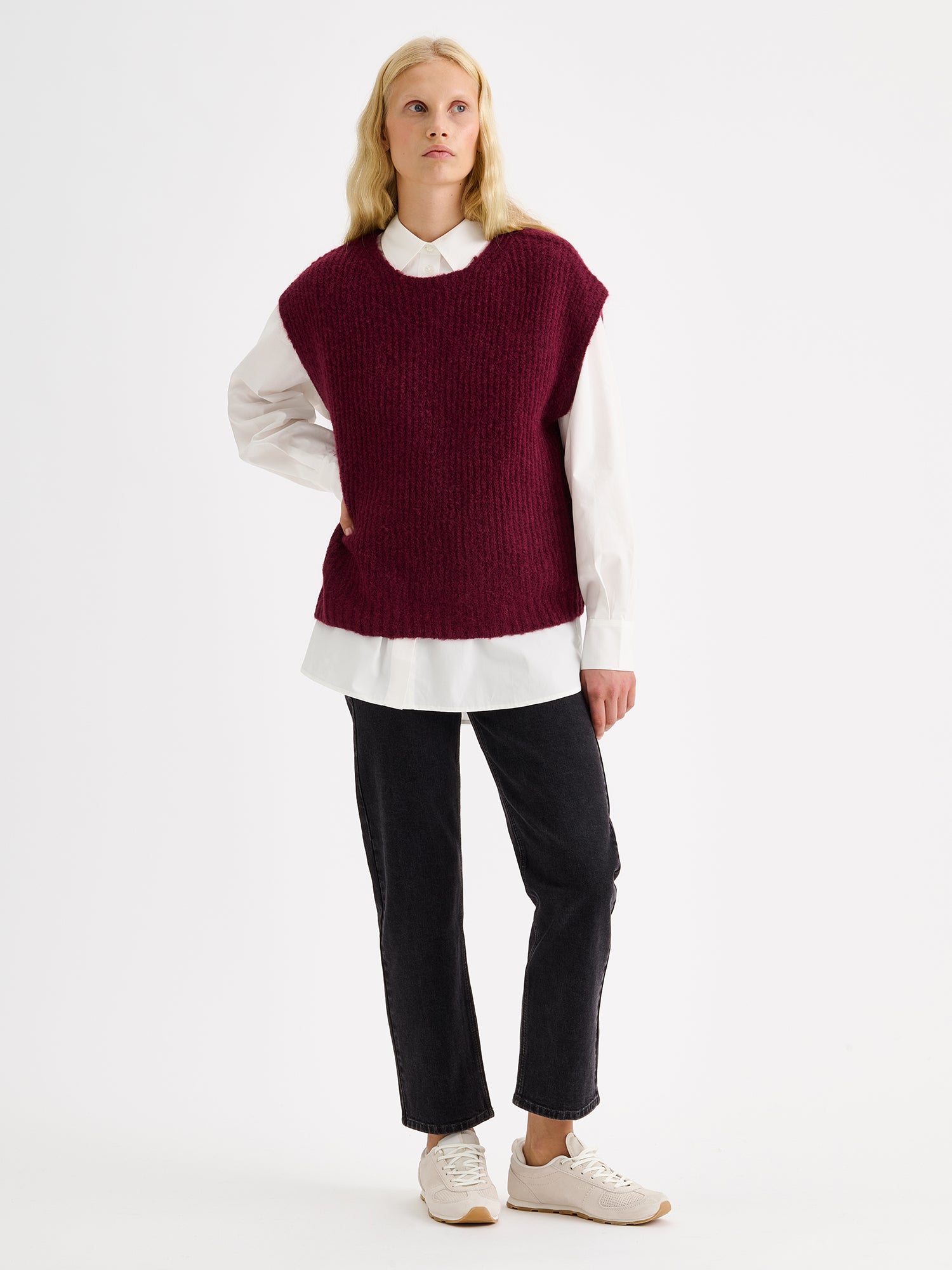 Nanso women's luxuriously soft and wonderfully chunky, the Karamelli knitted vest in a rich wine-red hue on a model with a white shirt and gray jeans