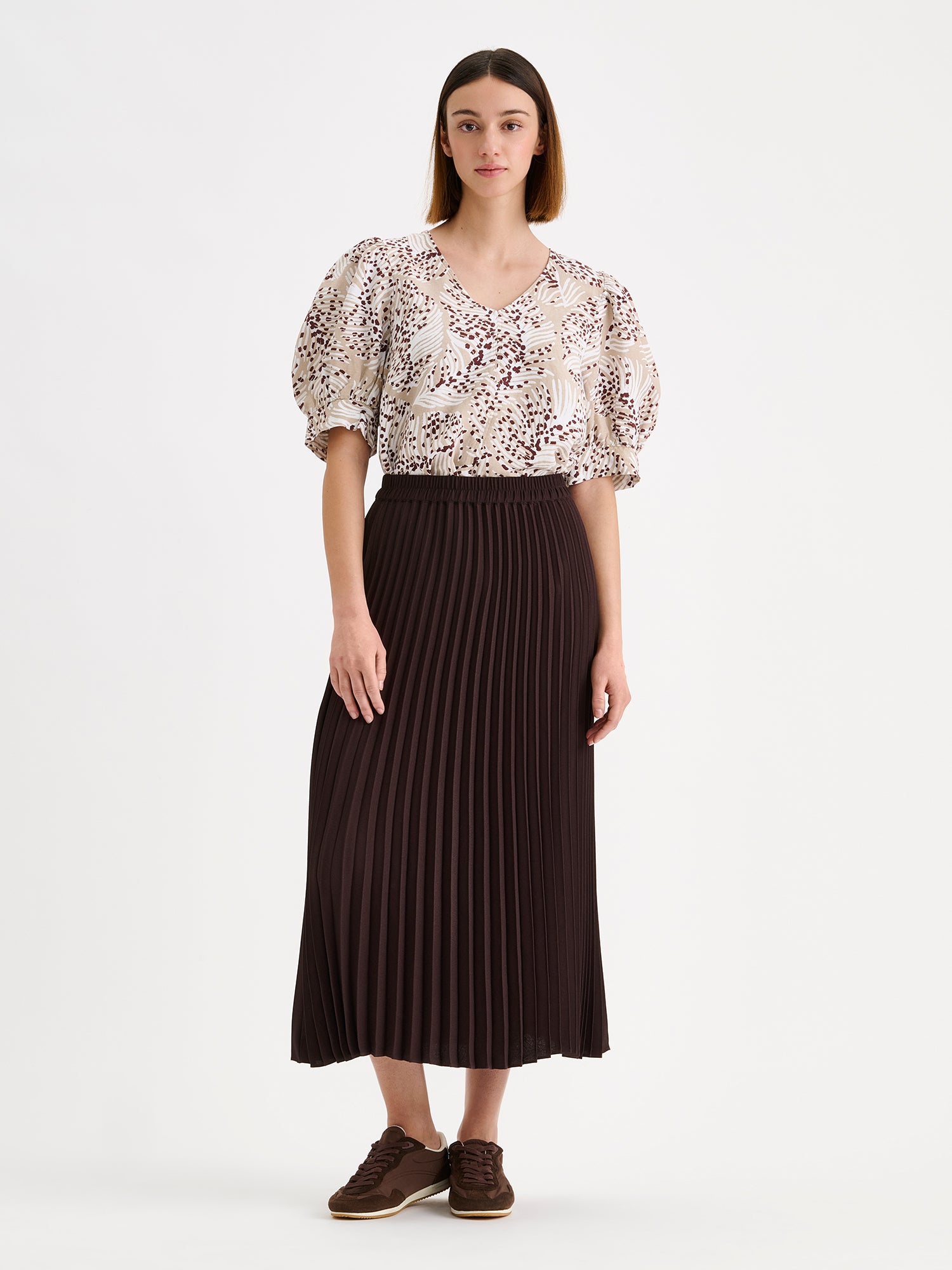 Nanso women's patterned Hilde blouse captivates with its elegant graphic and delicate pattern, flattering V-neckline and puff-sleeves. Pictured on a model with dark brown pleated midi skirt.