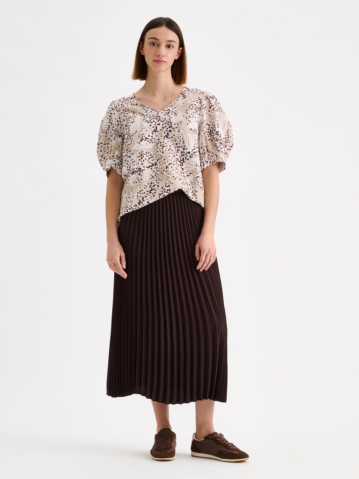 Nanso women's patterned Hilde blouse captivates with its elegant graphic and delicate pattern, flattering V-neckline and puff-sleeves. Pictured on a model with dark brown pleated midi skirt.