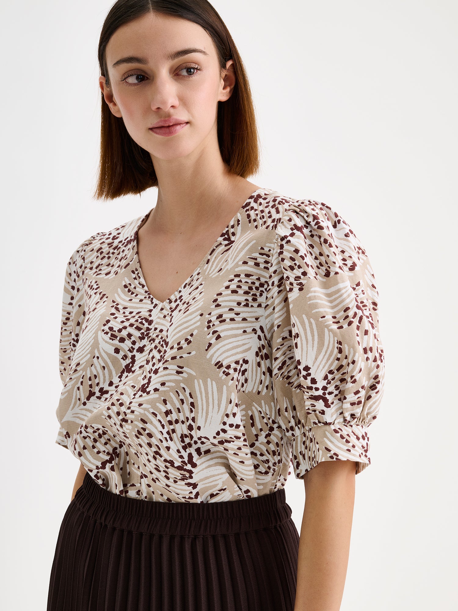 Nanso women's patterned Hilde blouse captivates with its elegant graphic and delicate pattern, flattering V-neckline and puff-sleeves. Pictured on a model with dark brown pleated midi skirt, close-up.