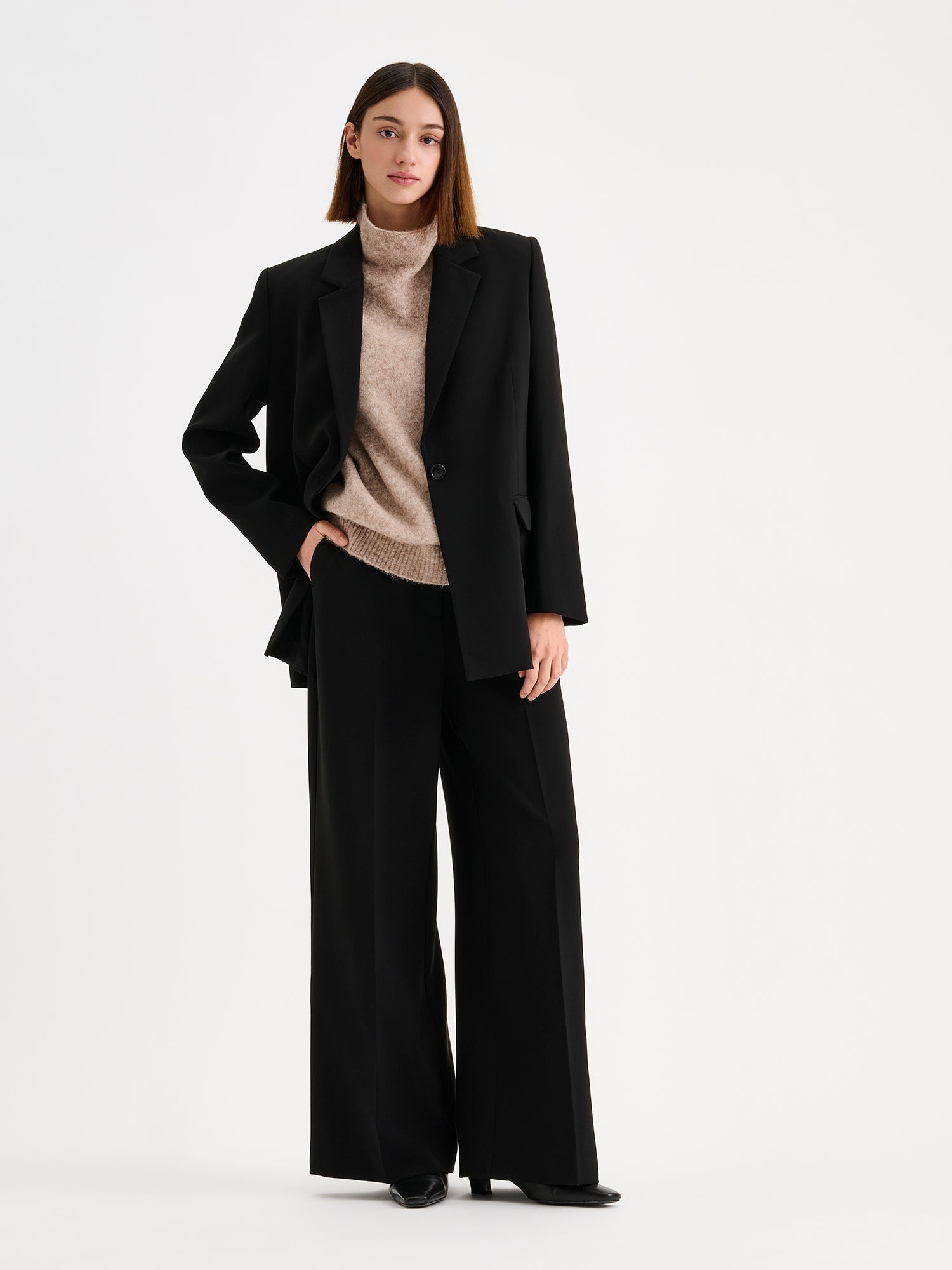 A stylish women's Eeva blazer in black with a relaxed yet structured silhouette, on a model with matching wide legged trousers and a beige knitted vest.