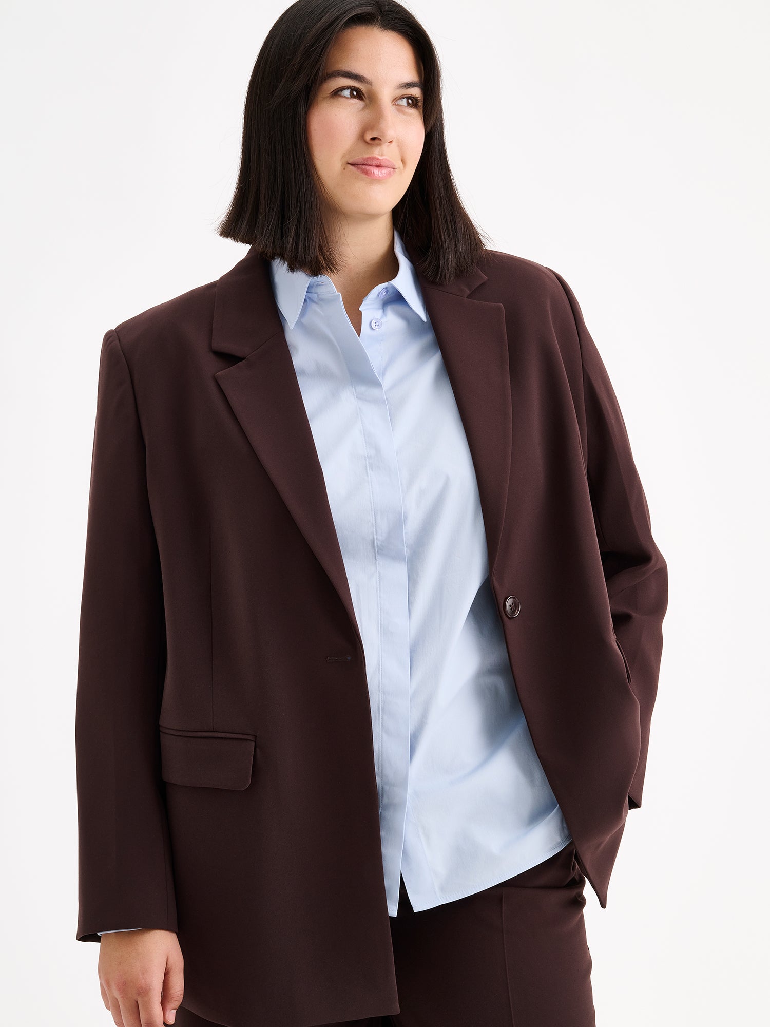 A stylish women's Eeva blazer in a rich brown shade with a relaxed yet structured silhouette, on a model with a light blue shirt