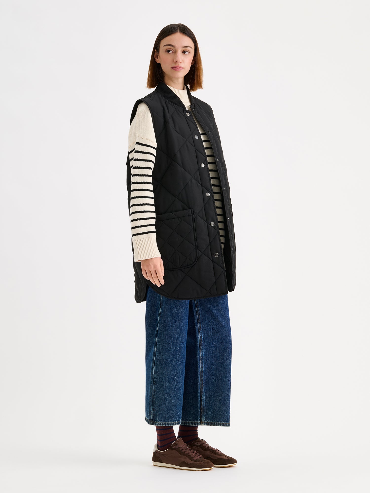 Nanso women's beautiful classic quilted vest in black, on a model with a striped sweater and wide blue jeans.
