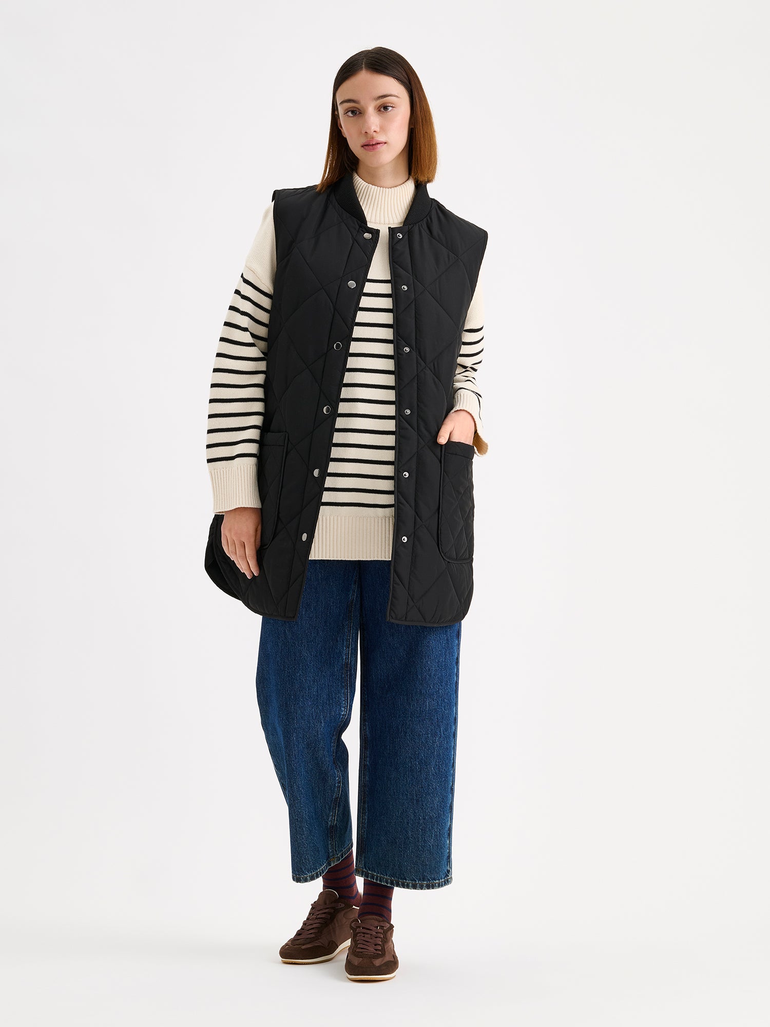 Nanso women's beautiful classic quilted vest in black, on a model with a striped sweater and wide blue jeans.