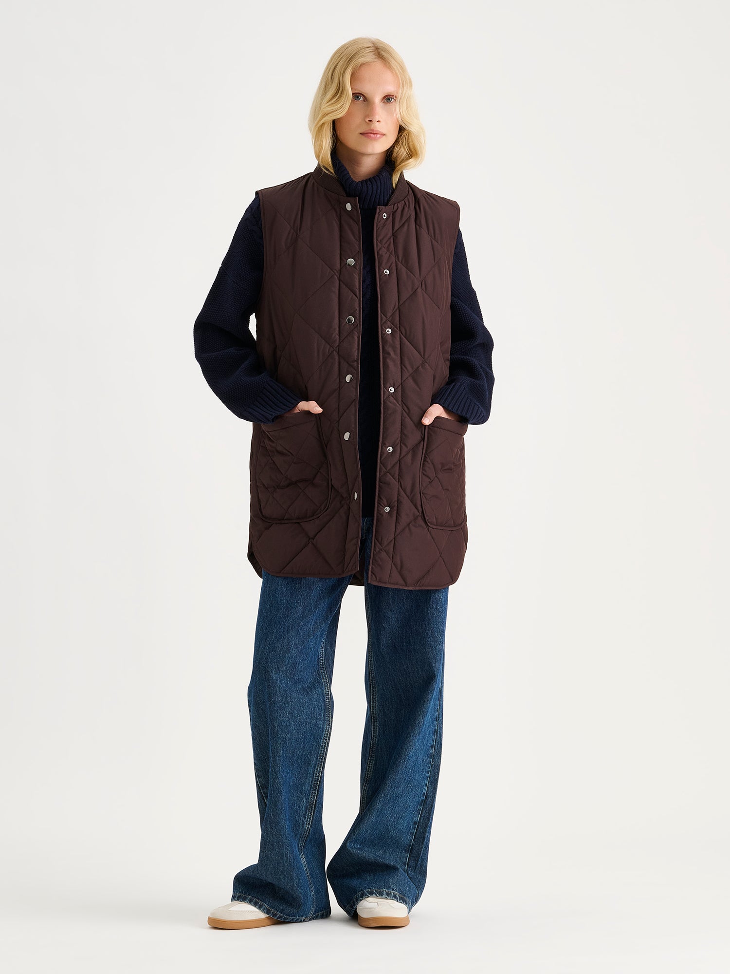 Nanso women's beautiful classic quilted vest in dark brown on a blonde model with a sweater and blue jeans. | S - 174cm